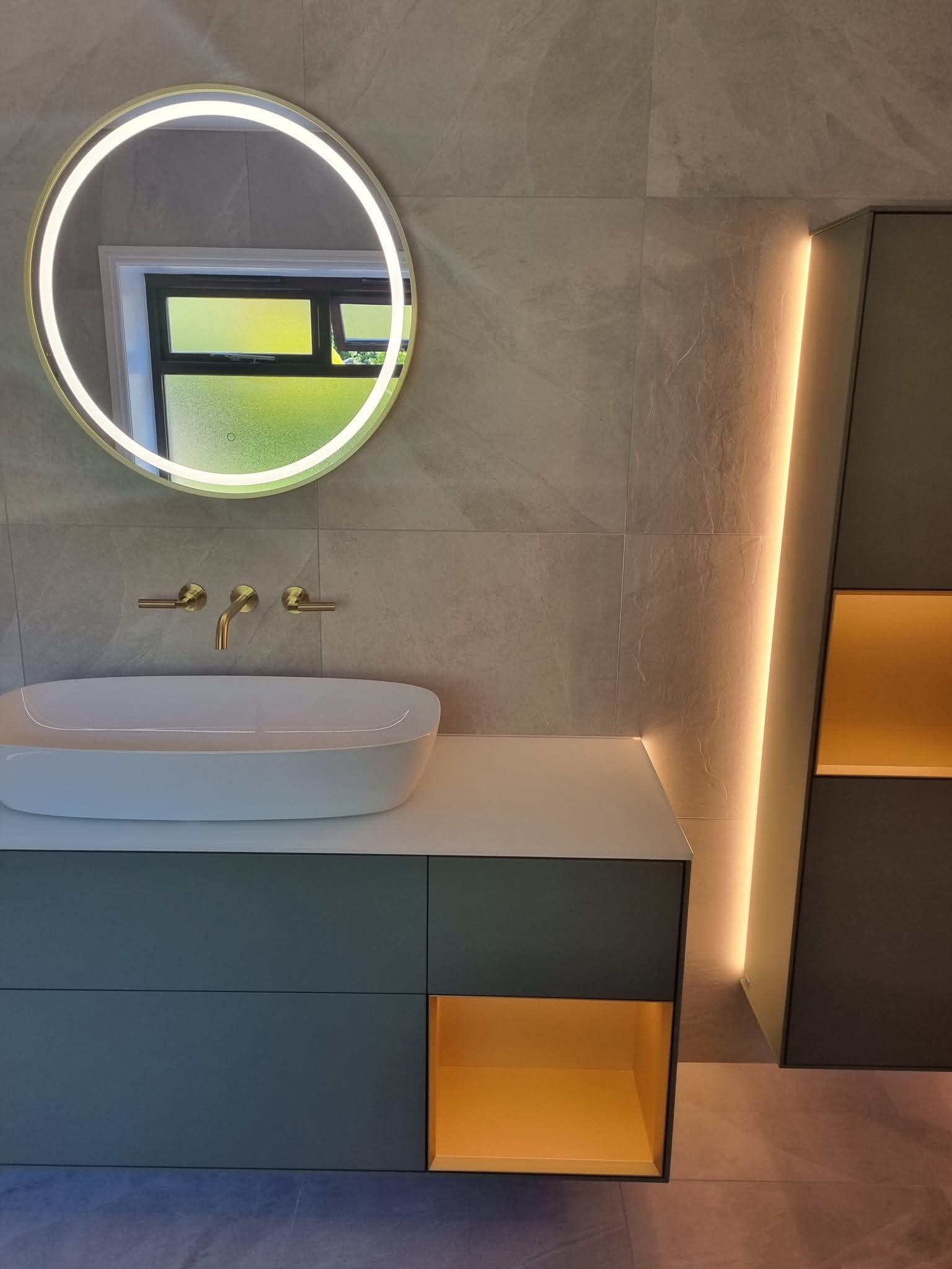 Modern bathroom vanity with blue-grey cabinetry, vessel basin, circular LED mirror and warm integrated lighting.