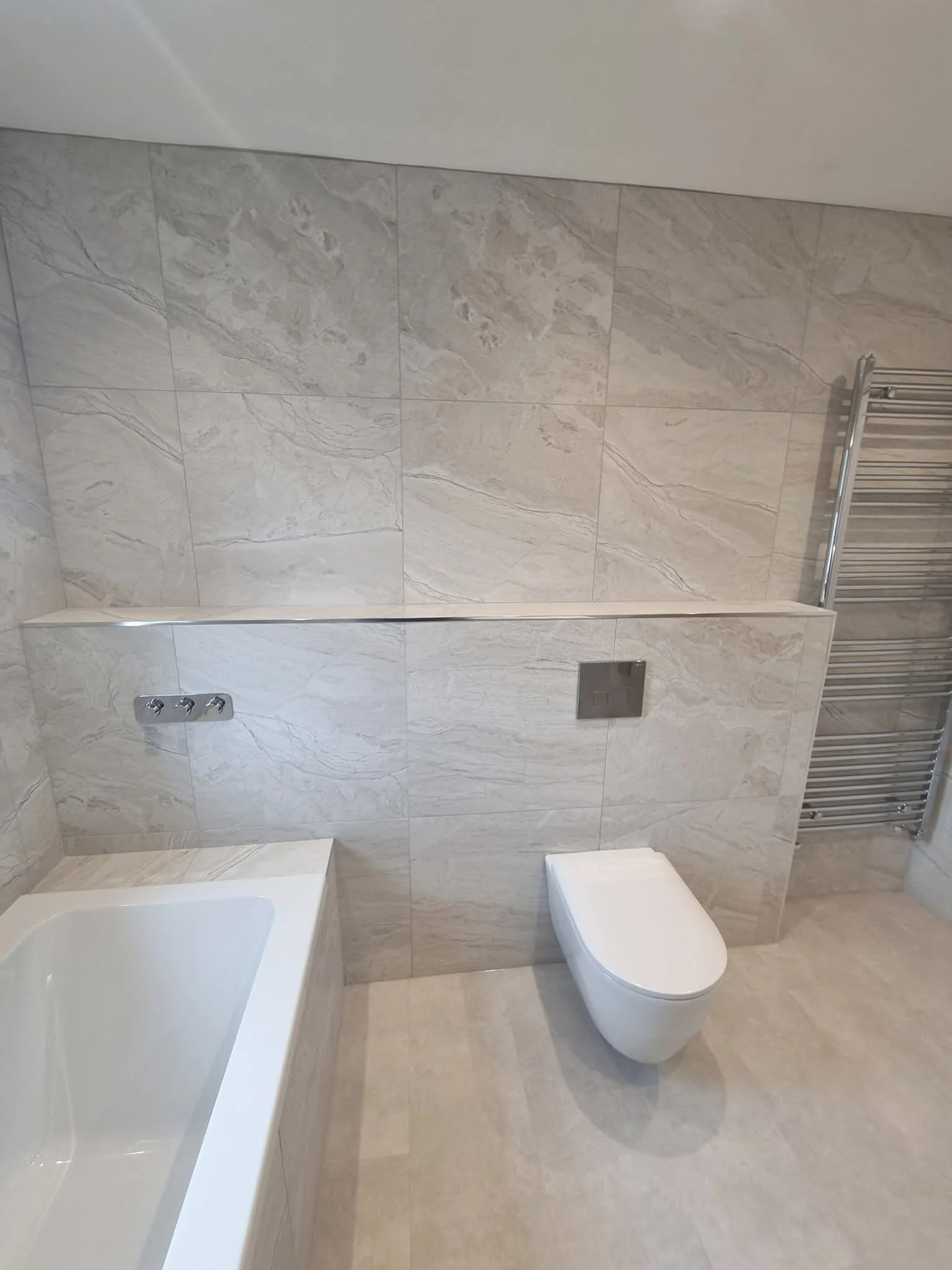 Light, modern family bathroom with fitted bathtub, wall-hung toilet and neutral stone-effect tiling.