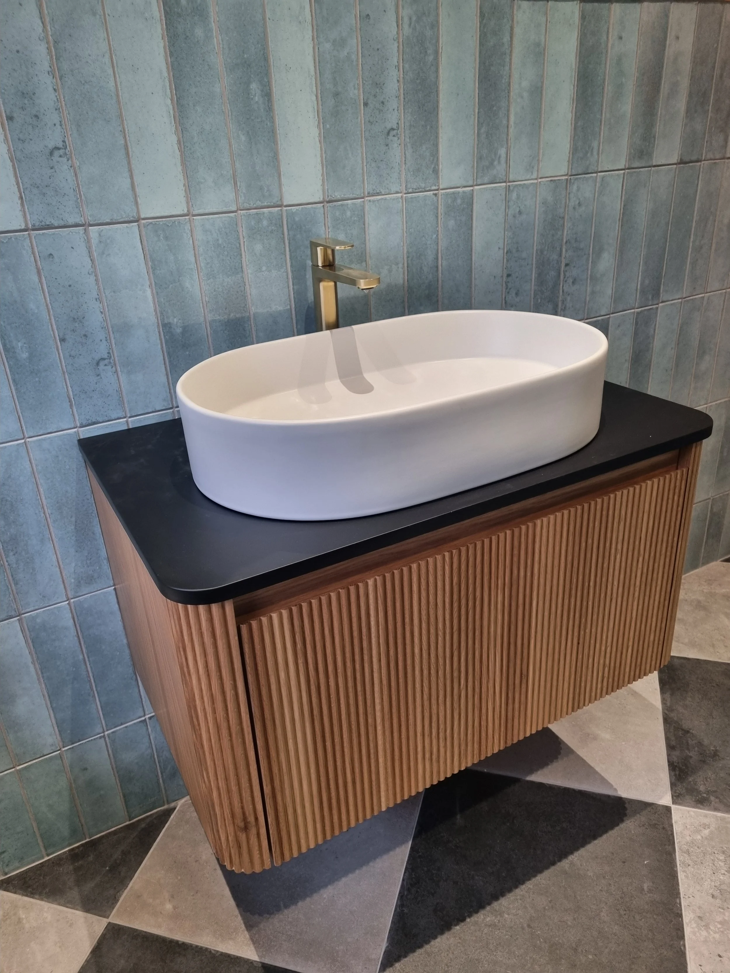 Modern bathroom vanity featuring a white countertop basin, brushed gold tap, fluted wood-effect cabinet and blue-green feature wall tiles.