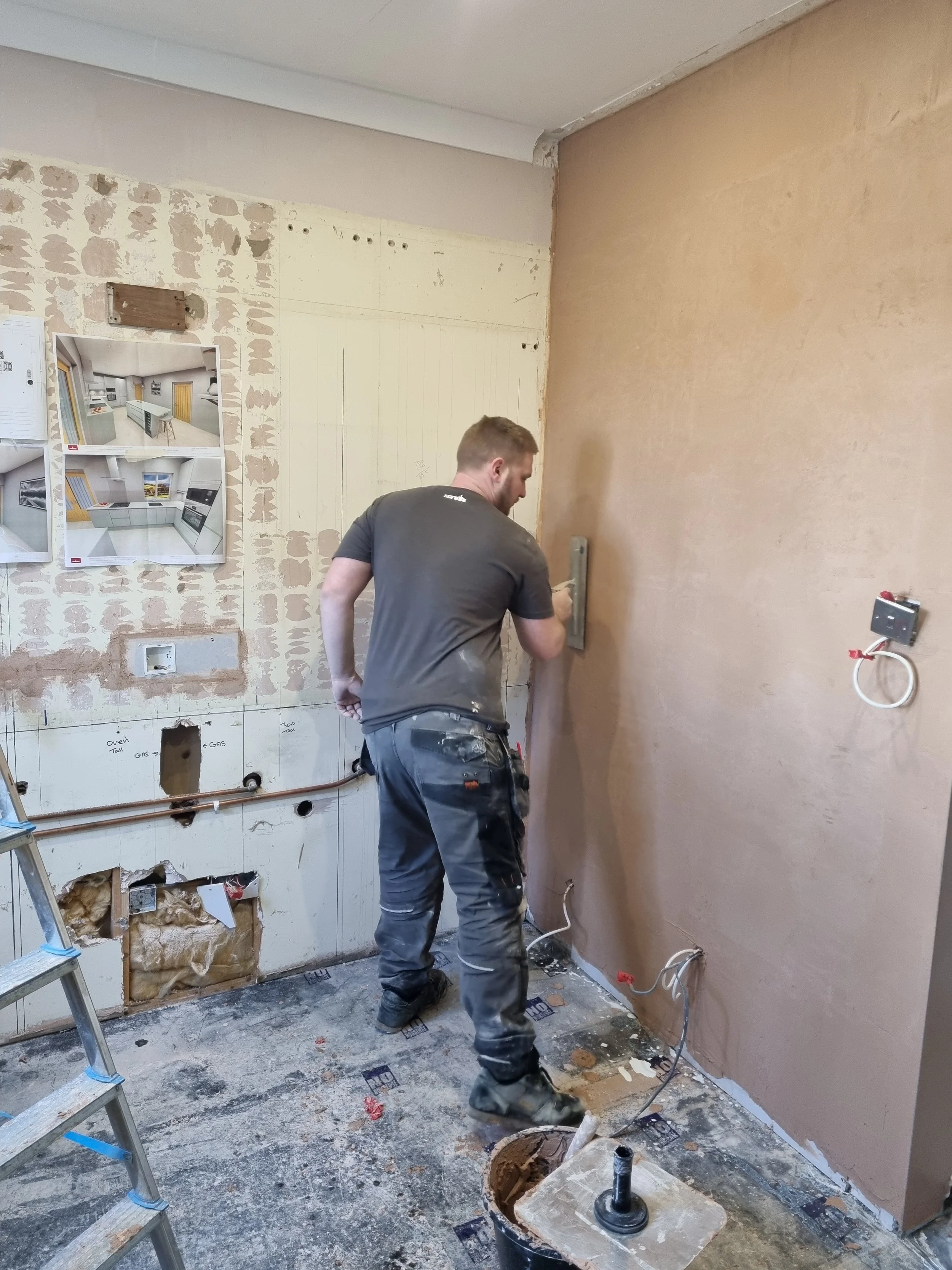 A man in work clothes is installing or repairing electrical wiring on a brown wall in a room under renovation. There is a ladder on the left side, and the floor is scattered with debris, tools, and construction materials.