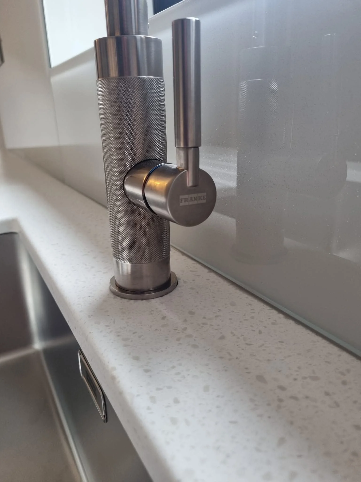 Close-up of a contemporary brushed metal kitchen mixer tap against a sleek glass splashback and light-coloured worktop.