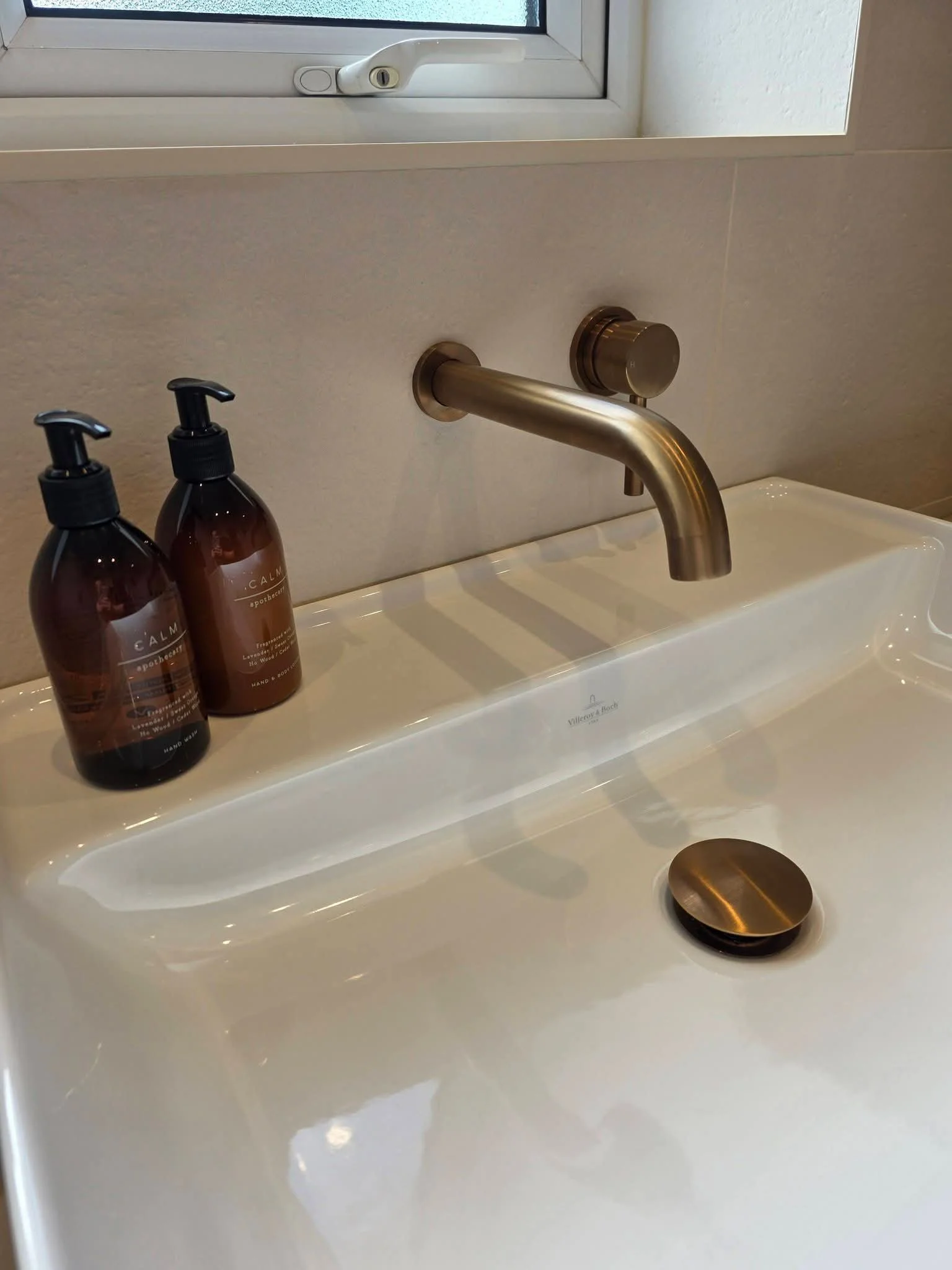 Contemporary white bathroom basin with wall-mounted brushed bronze tap, matching waste and neutral stone-effect tiles.