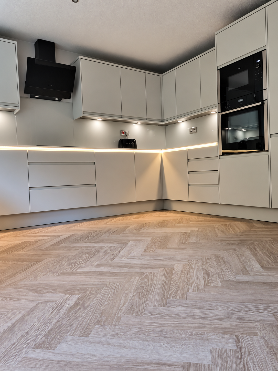 Modern handleless kitchen renovation by Oliver Francis Interiors, featuring matt dove-grey cabinetry, integrated appliances, glass splashbacks, LED lighting and herringbone flooring.