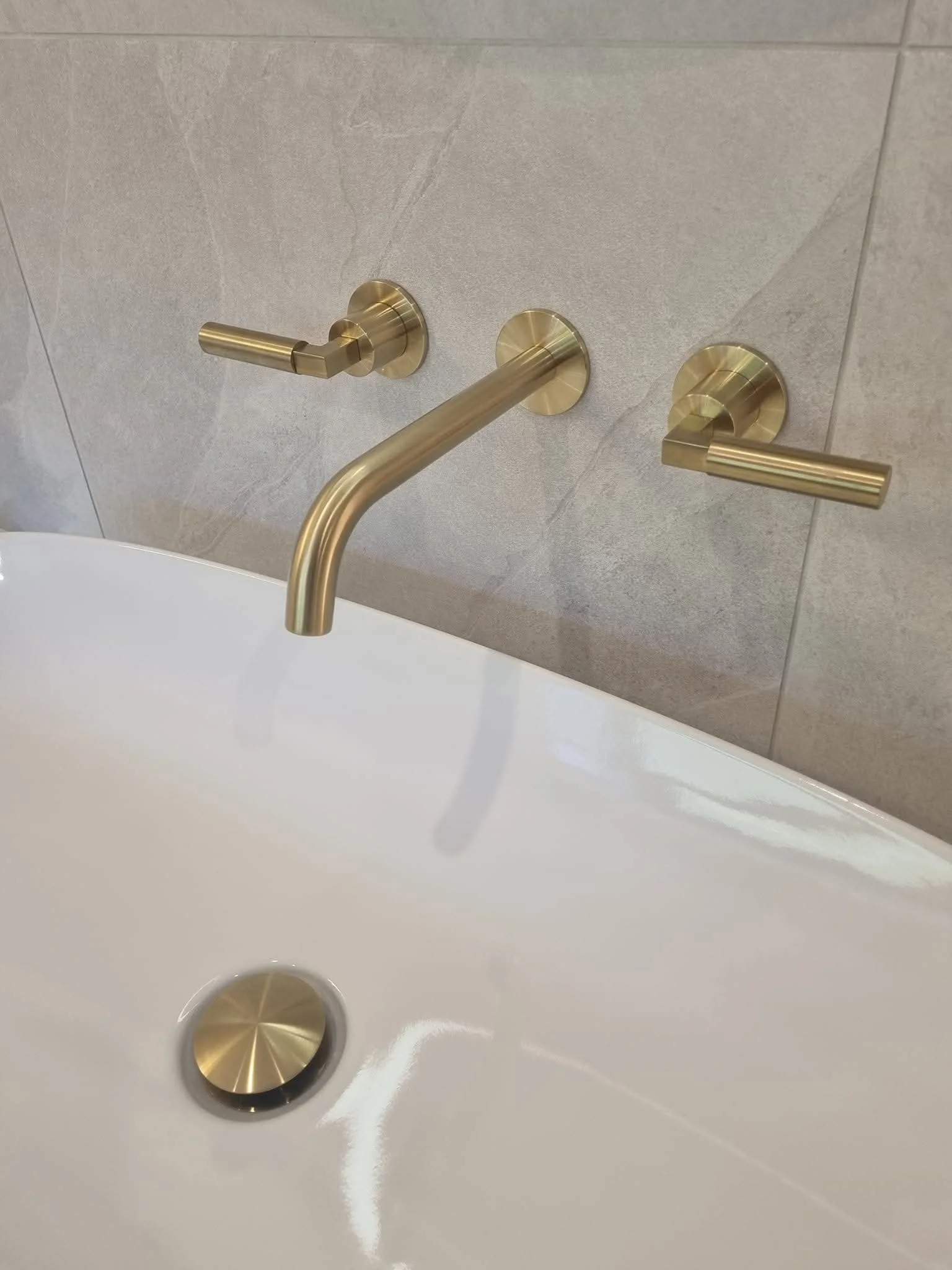 Close-up of brushed gold wall-mounted bath tap and matching controls above a contemporary white bathtub.