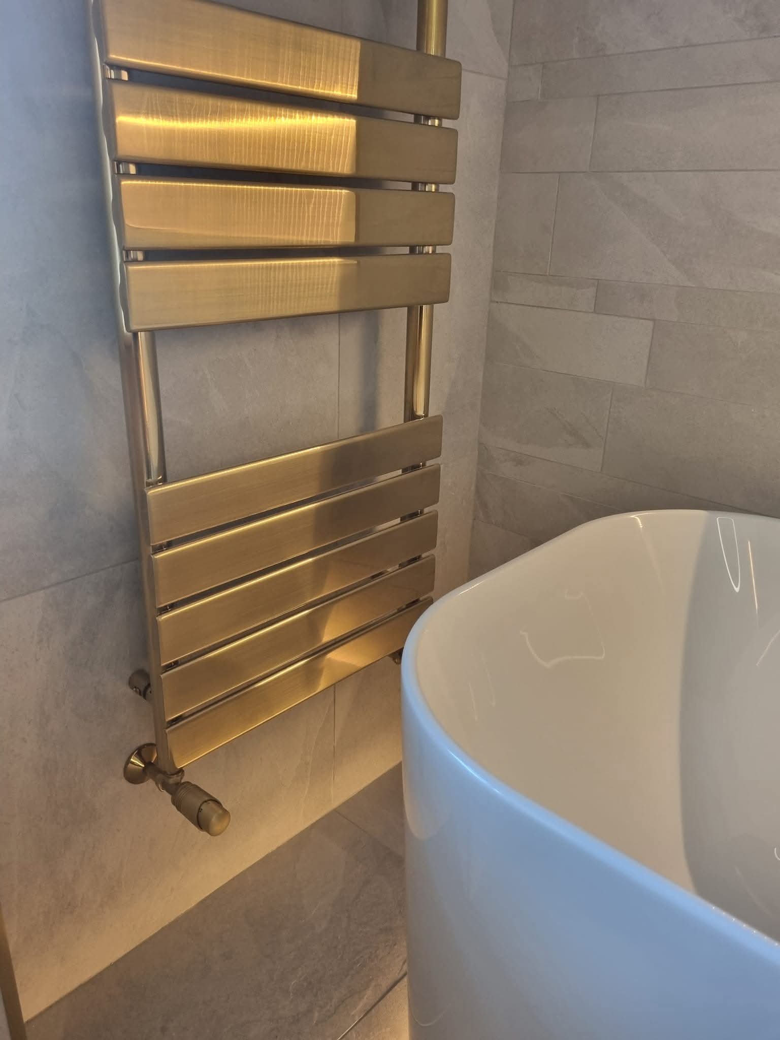 Brushed gold heated towel rail mounted on neutral tiled bathroom wall beside a freestanding bath.