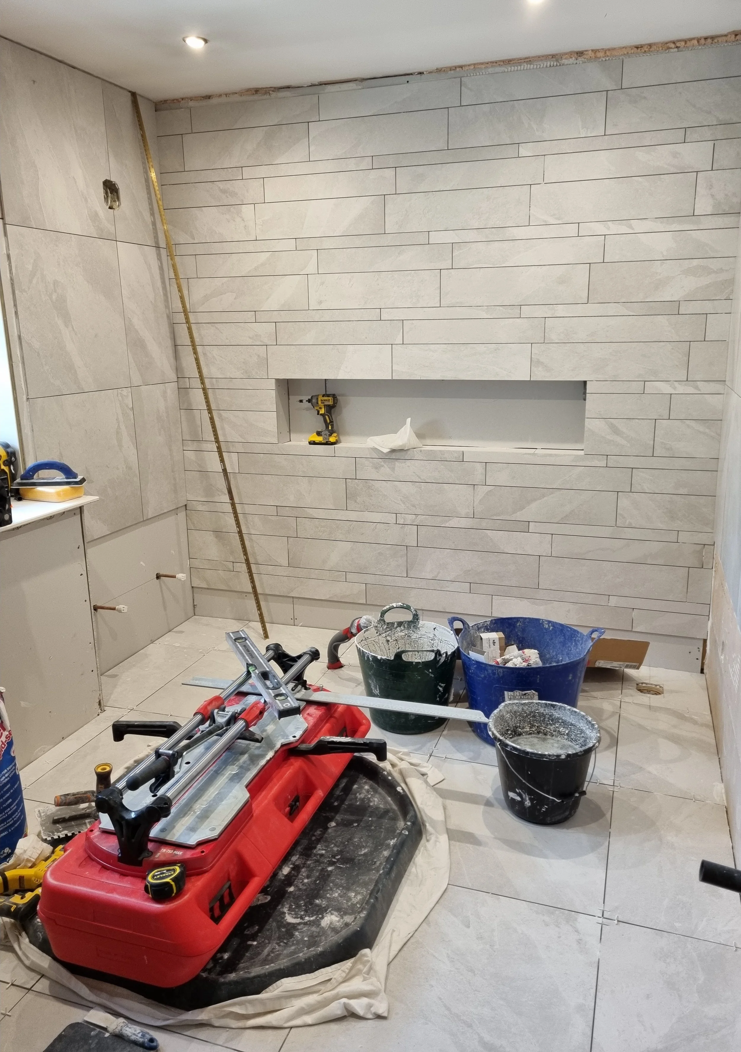 Interior of a room under construction with partially tiled walls and various tools and construction materials on the floor, including a red toolbox, a cordless drill, buckets, and a level.