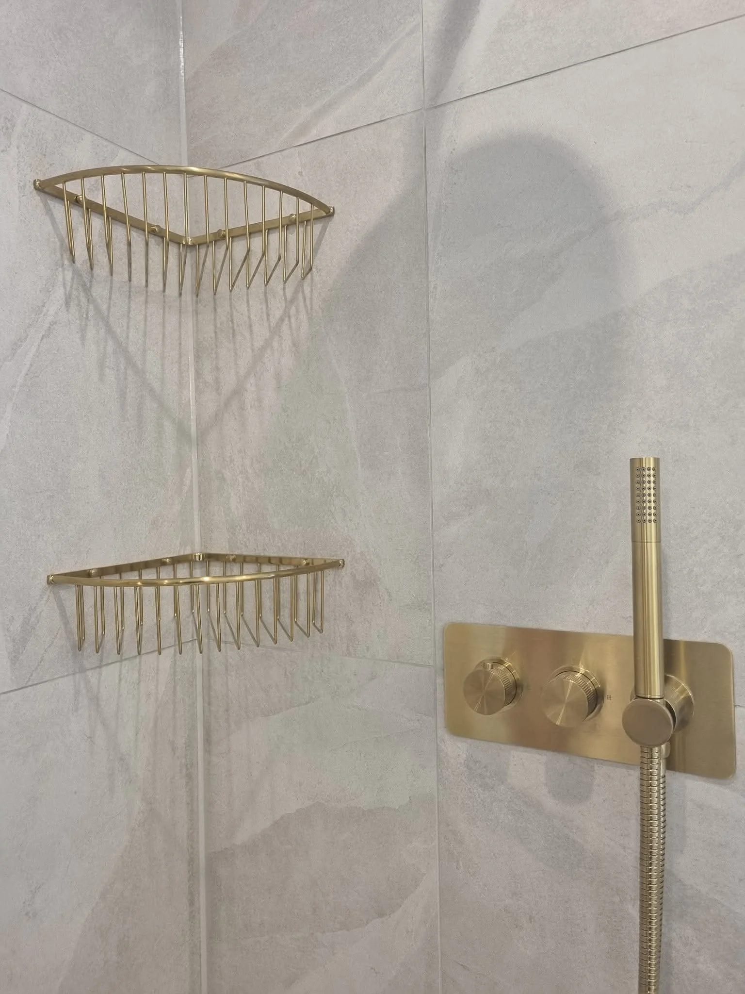 Brushed gold shower controls and corner storage baskets against light stone-effect bathroom tiles.