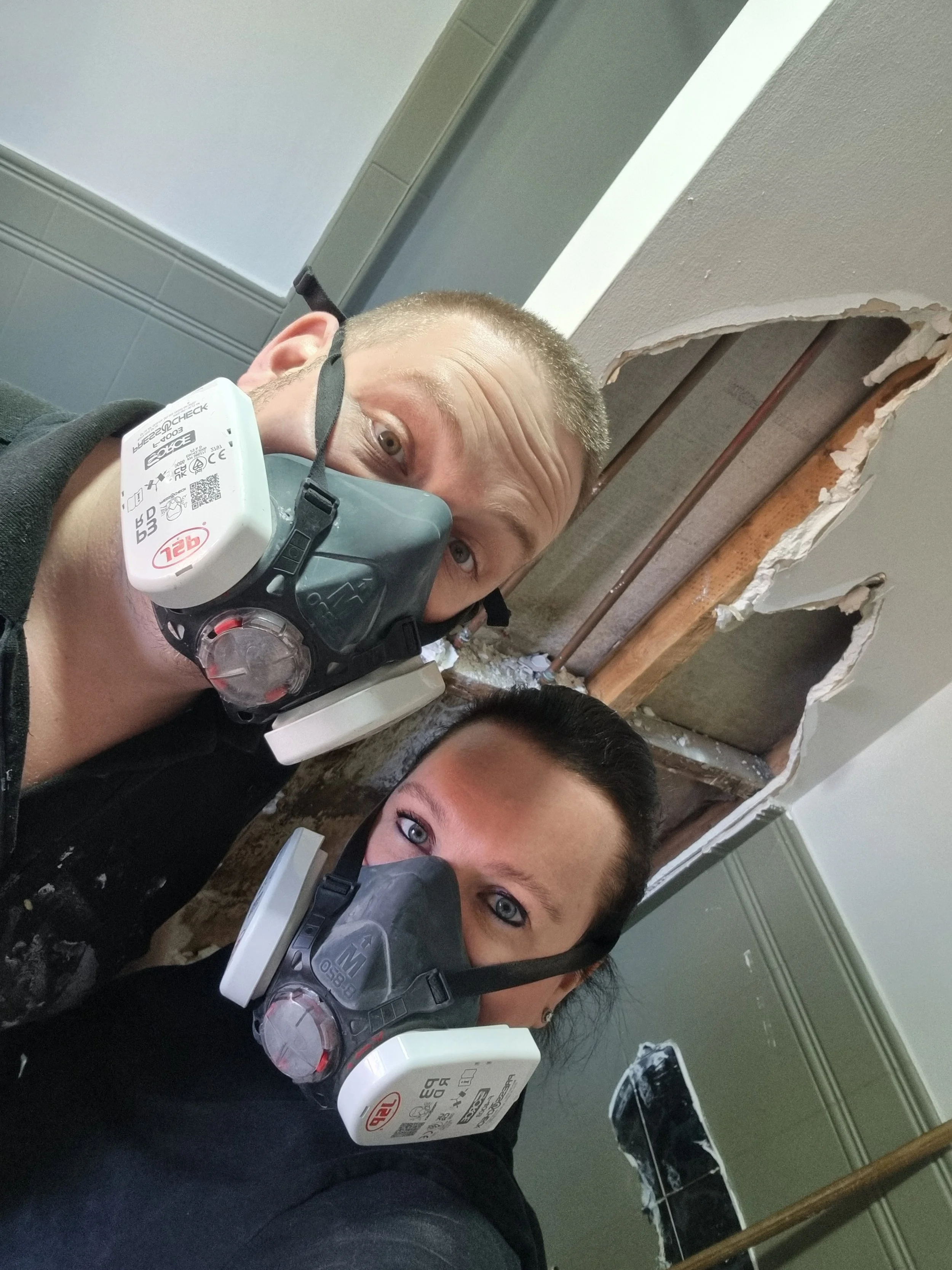 Two people wearing masks with air filters attached to their faces, working on a ceiling with a large hole exposing pipes and drywall. The ceiling is partially demolished.