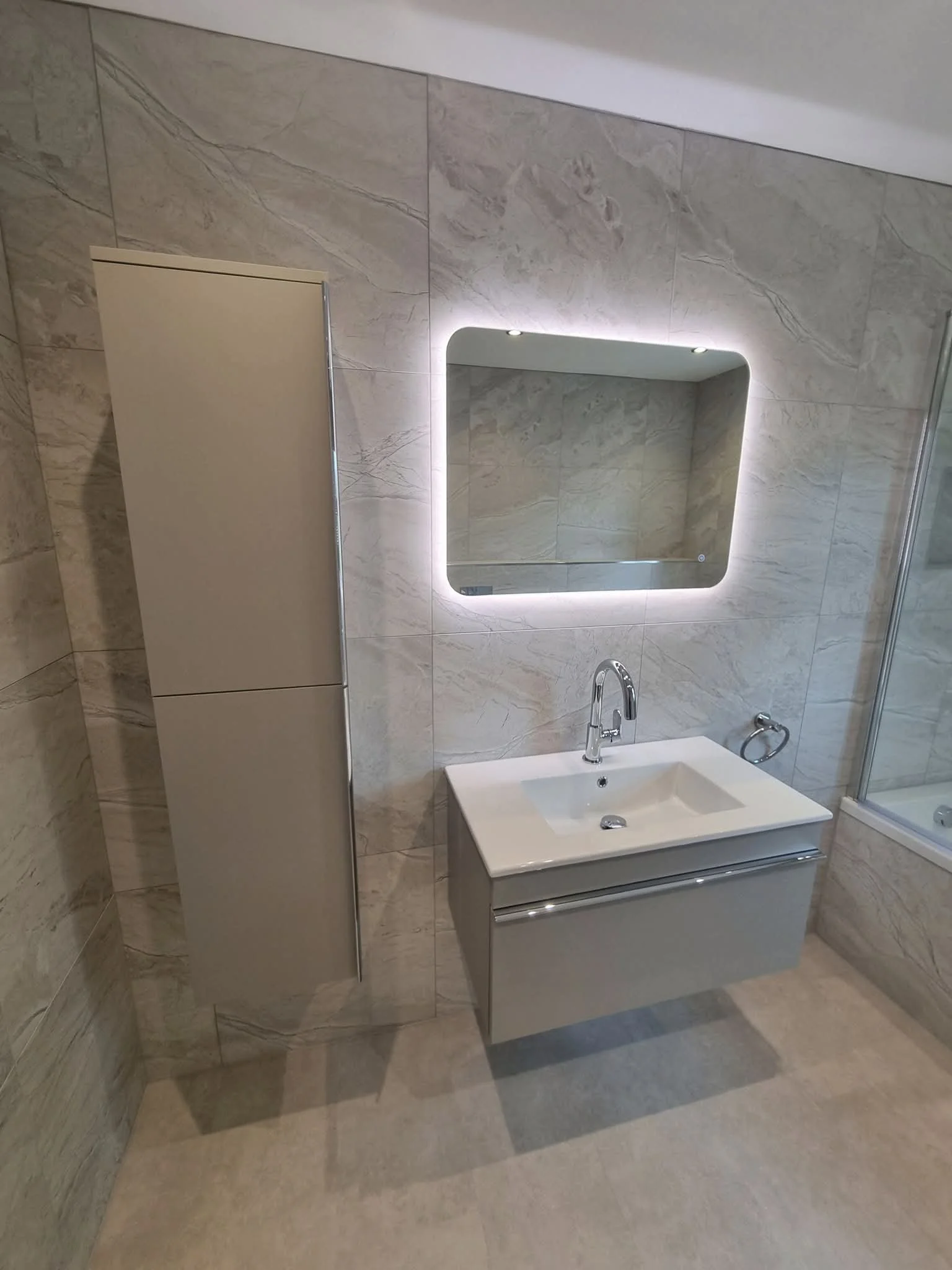 A bathroom with beige marble walls and floor, a backlit rectangular mirror, a white vanity with a sink and faucet, a tall beige cabinet, and a towel ring on the wall.