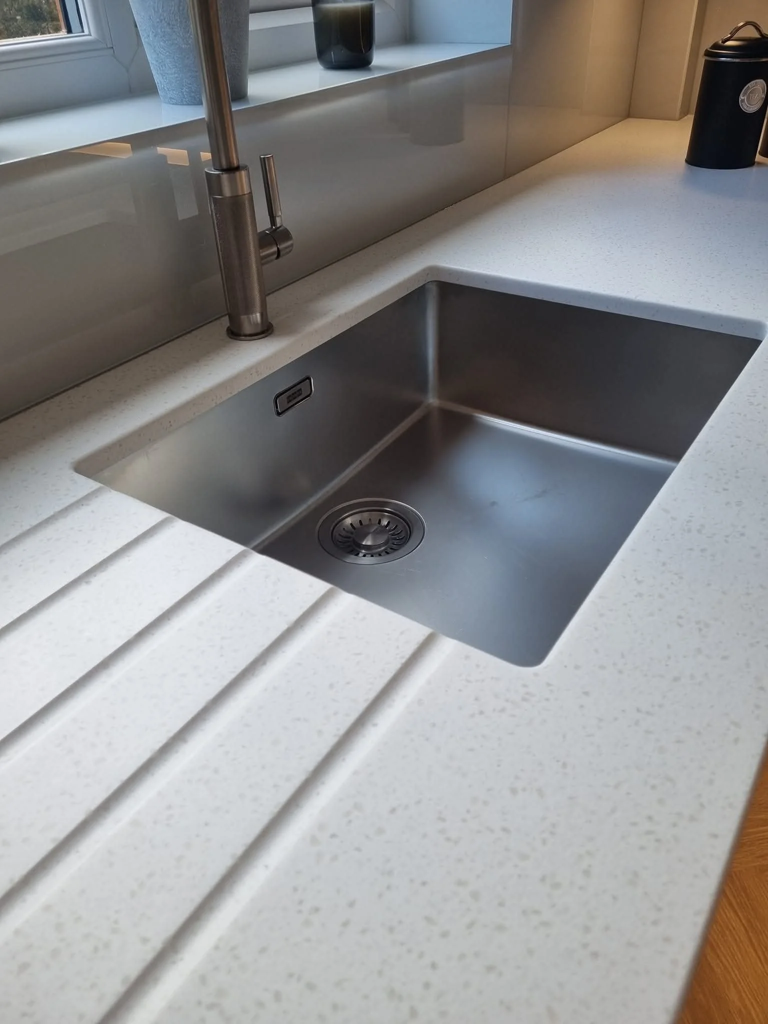 Close-up image of a kitchen sink with a modern metallic faucet, set in a white speckled countertop with a built-in draining board, next to a window with potted plants and a black container on the counter.