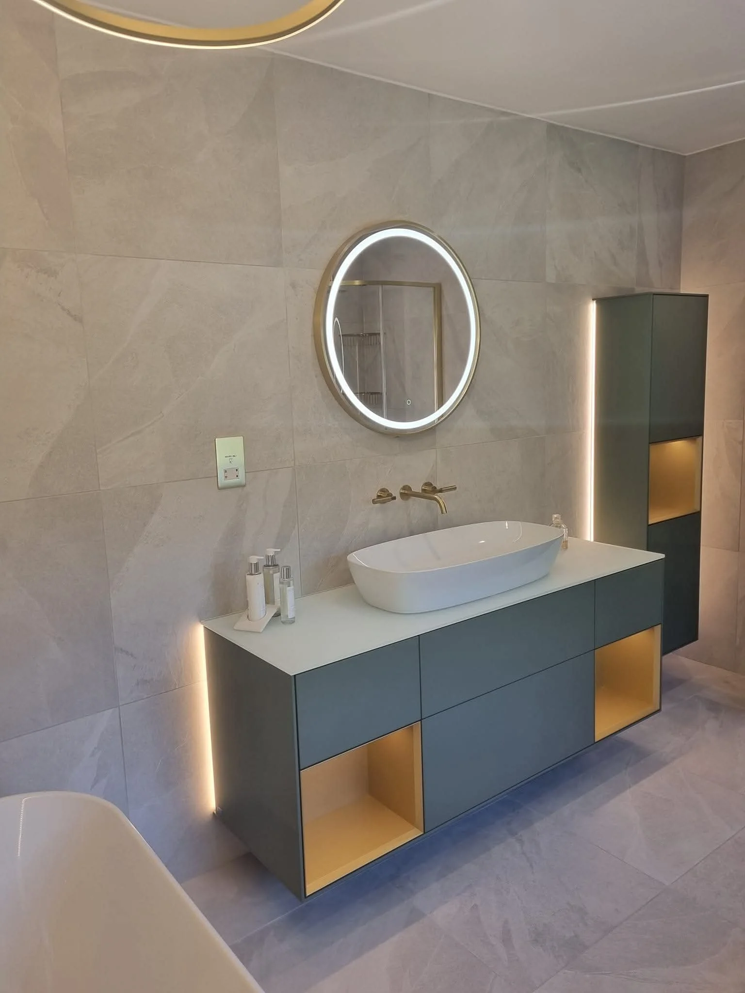 Modern bathroom vanity with a rectangular vessel sink, a round illuminated mirror, a wall cabinet with lighting accents, and toiletry bottles on the countertop. The wall is tiled with large neutral-colored tiles.