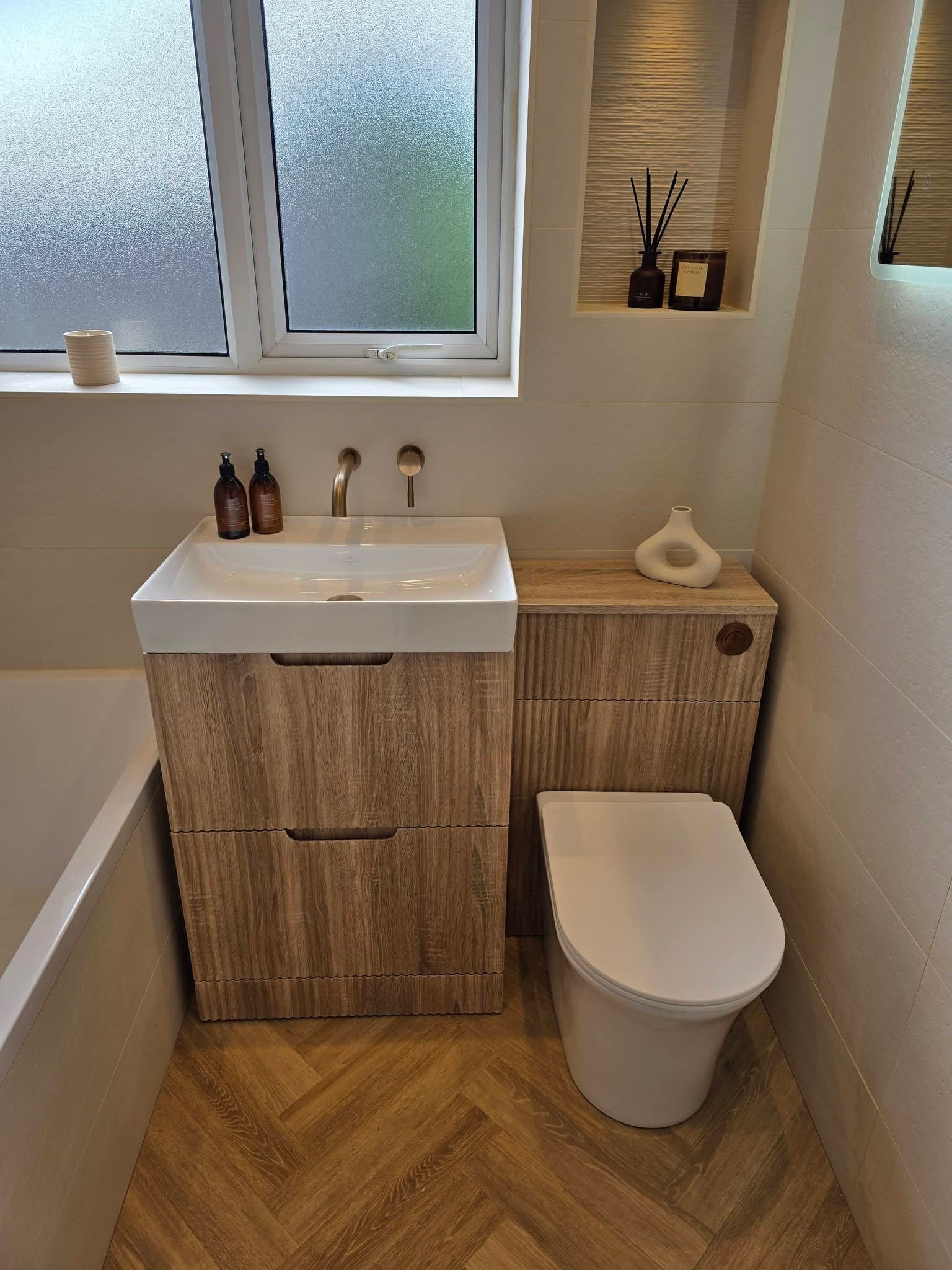 A modern bathroom with a white rectangular sink, a wooden vanity, a frosted window, and a white toilet, with decorative bottles and reed diffusers.