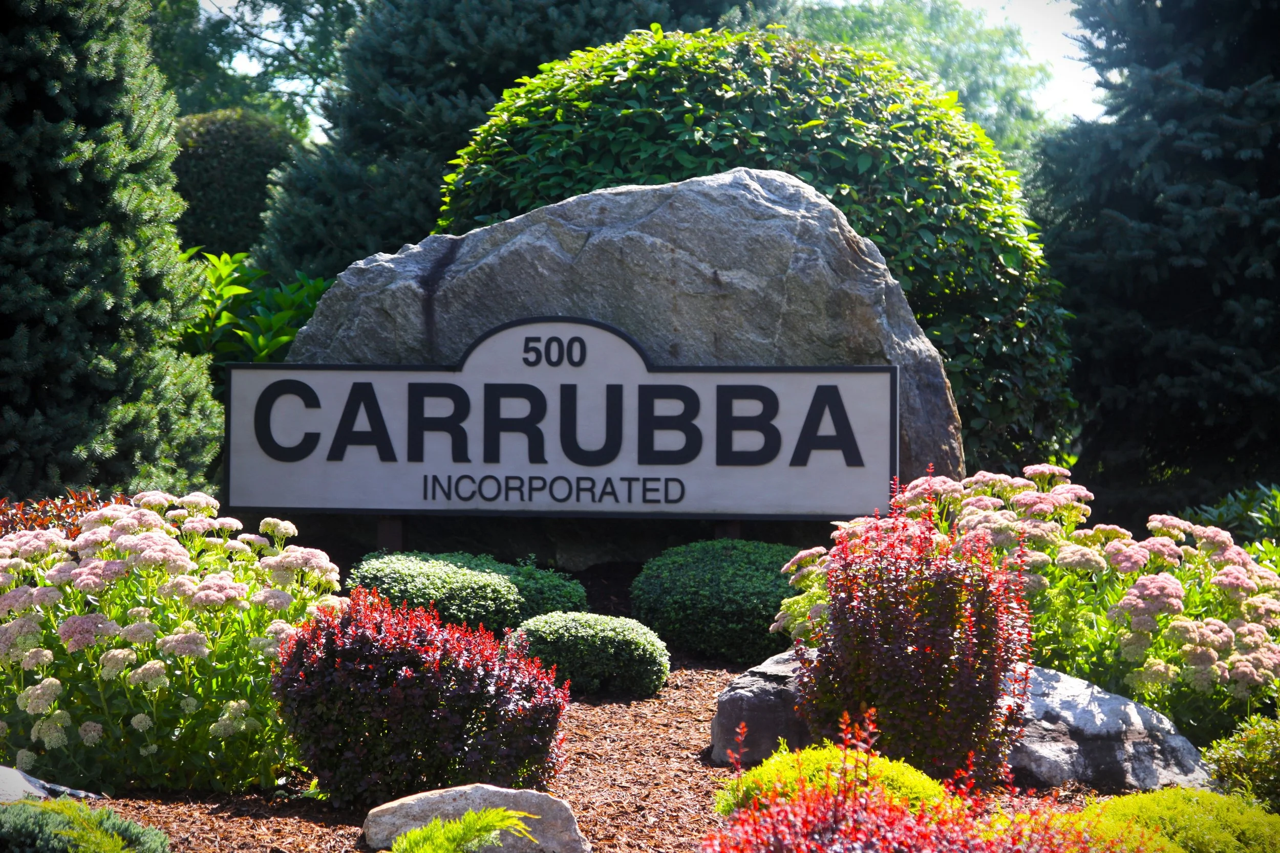 Sign with the text '500 Carruba Incorporated' surrounded by a landscaped garden with various green shrubs, pink, red, and white flowers, and large rocks, with trees and blue sky in the background.