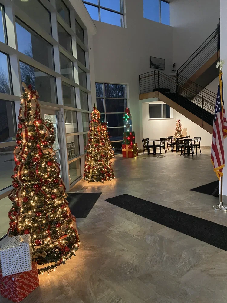 Decorated Christmas trees with lights and ornaments inside a modern building with large windows, a staircase, and an American flag.