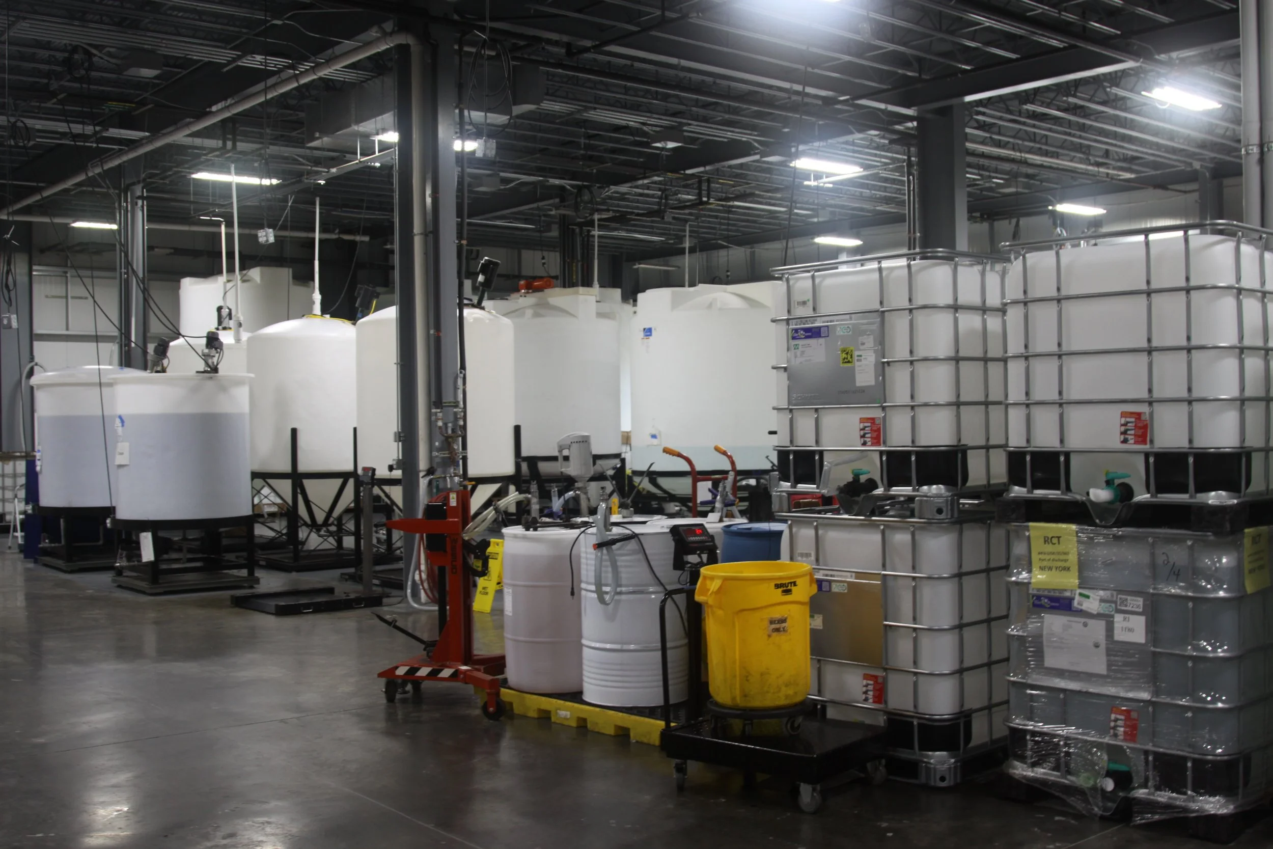 Industrial warehouse with large white plastic tanks, metal shelving units, yellow and black safety barrels, and various equipment and piping.