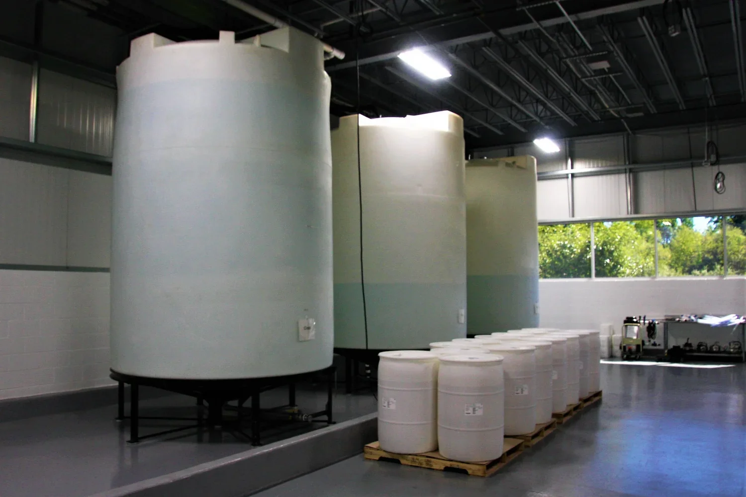 Large white storage tanks and drums inside an industrial building with a window showing greenery outside.