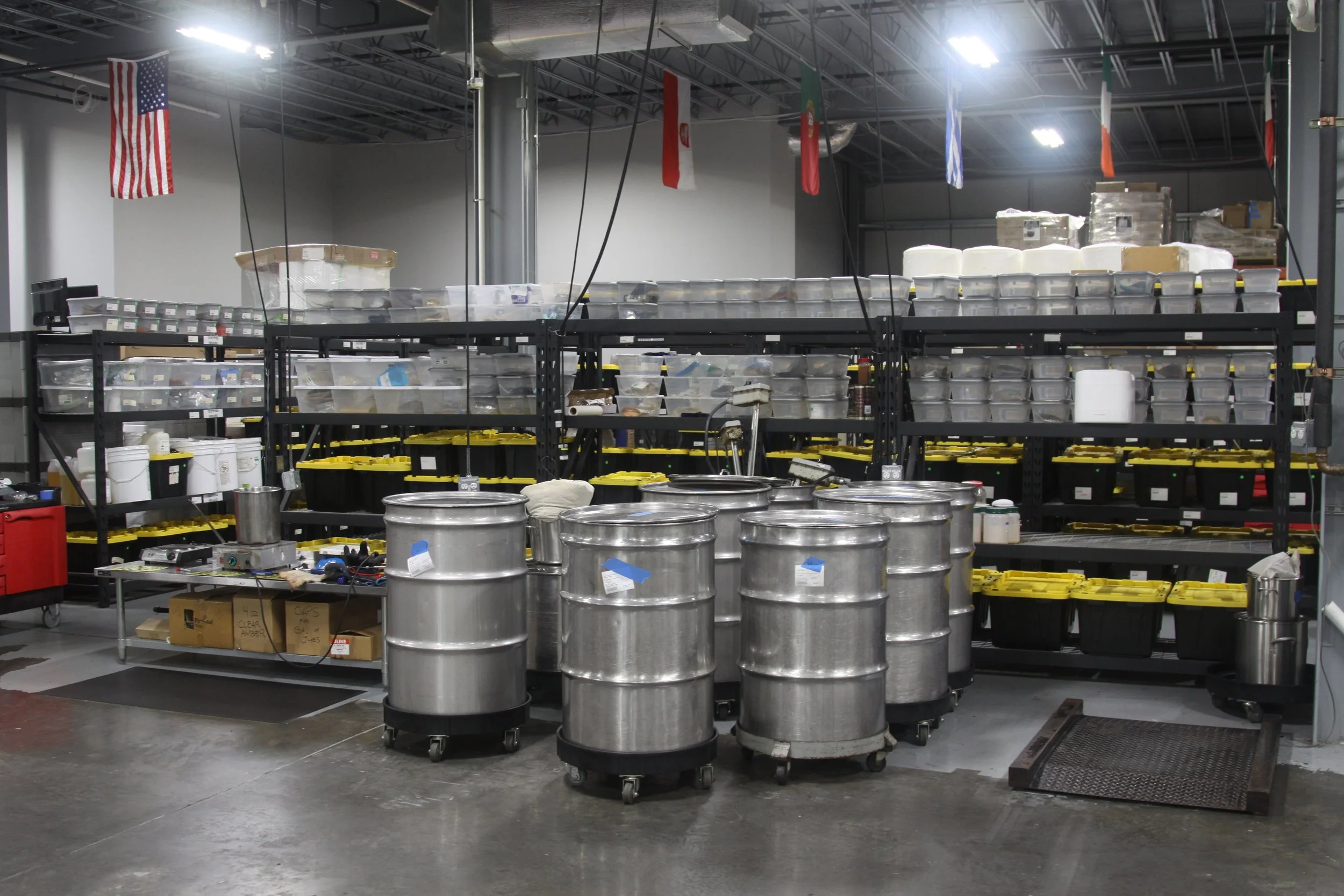Warehouse storage area with metal shelves holding plastic containers, cardboard boxes, and large metal drums on a concrete floor.