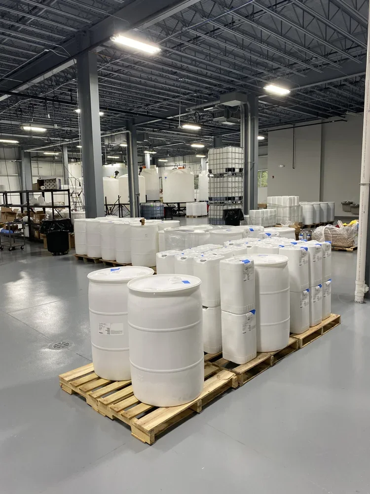 Large warehouse with industrial lighting and metal ceiling, containing pallets of white plastic drums and containers, some with blue caps, arranged on wooden pallets.