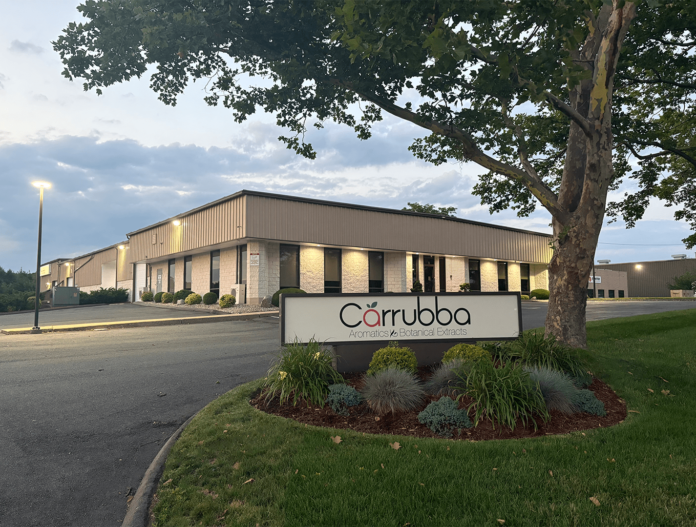 Photo of a commercial building with a sign in front that reads 'Carrubba Aromatics & Botanical Extracts.' The building has a beige and gray exterior with windows, surrounded by landscaped bushes and a tree, under a cloudy sky during dusk.