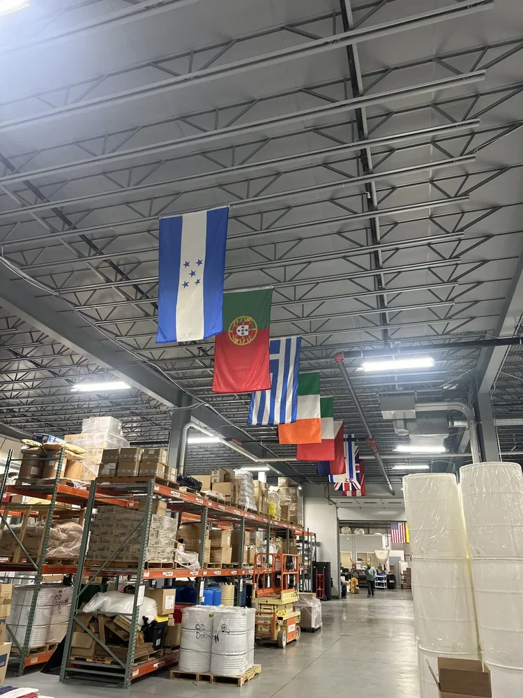 Warehouse aisle with shelves full of boxes and supplies, multiple national flags hanging from the ceiling, including Greece, Portugal, and Greece again, and a forklift and packing materials visible.