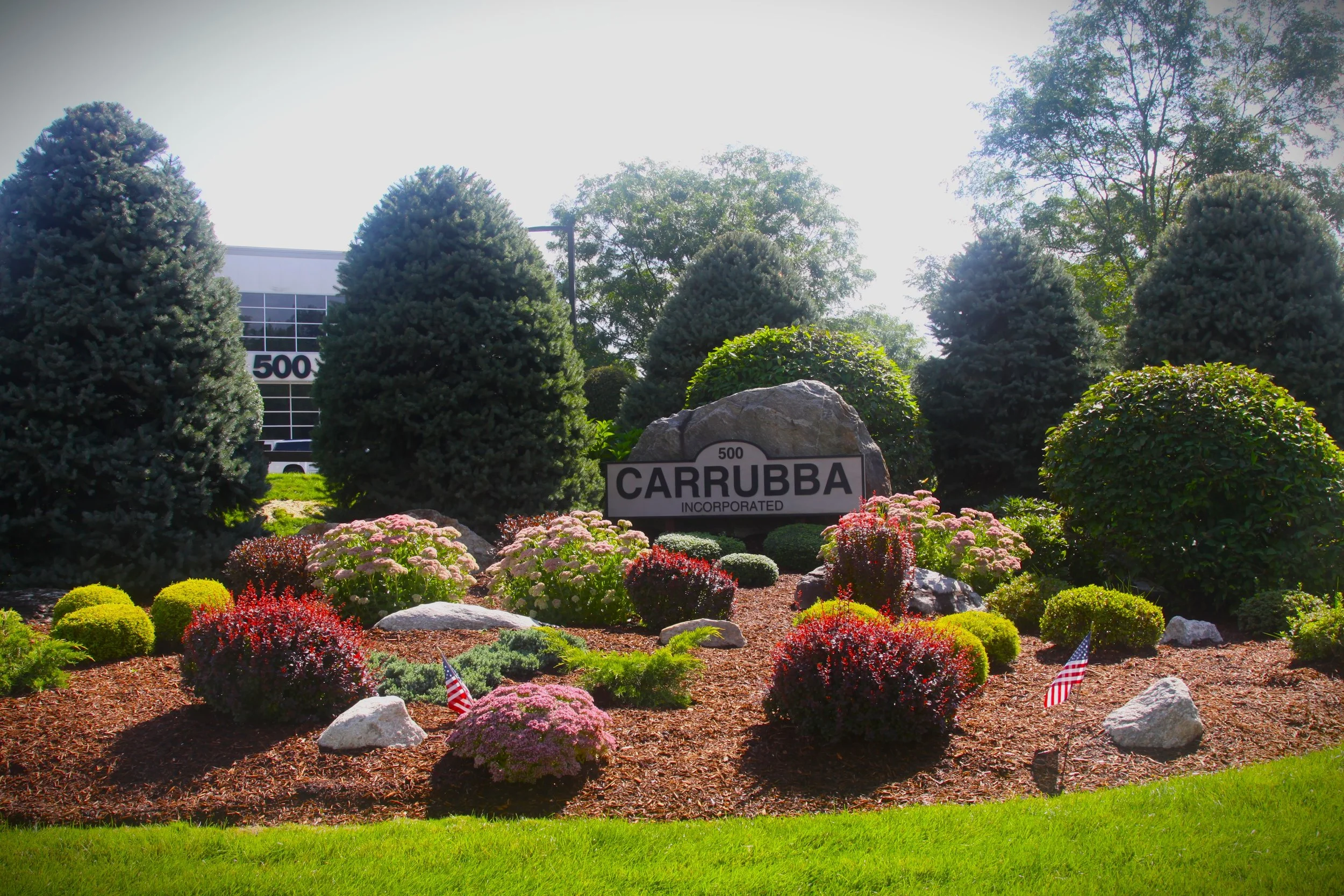 A landscaped area with a large rock in front of a sign that reads "500 CARRUBBA INCORPORATED." The area features colorful flowers and shrubs, with tall pine trees and leafy trees in the background. American flags are placed among the flowers.