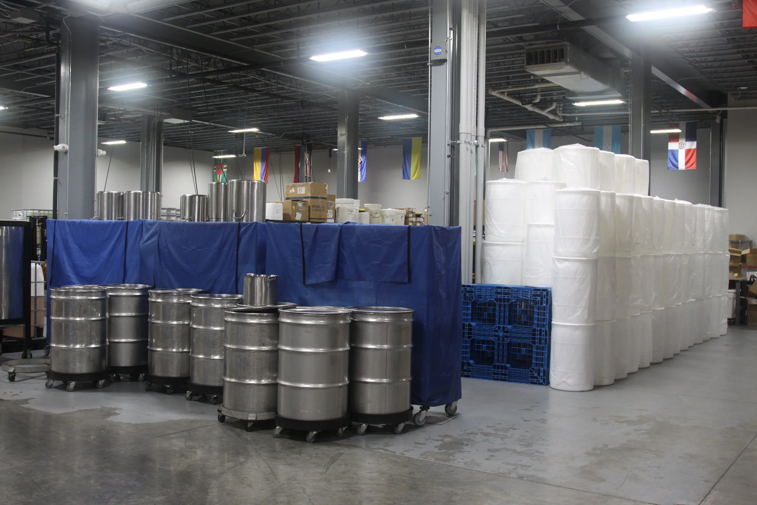 Industrial warehouse with metal barrels, large rolls of plastic or fabric, and flags hanging from the ceiling.