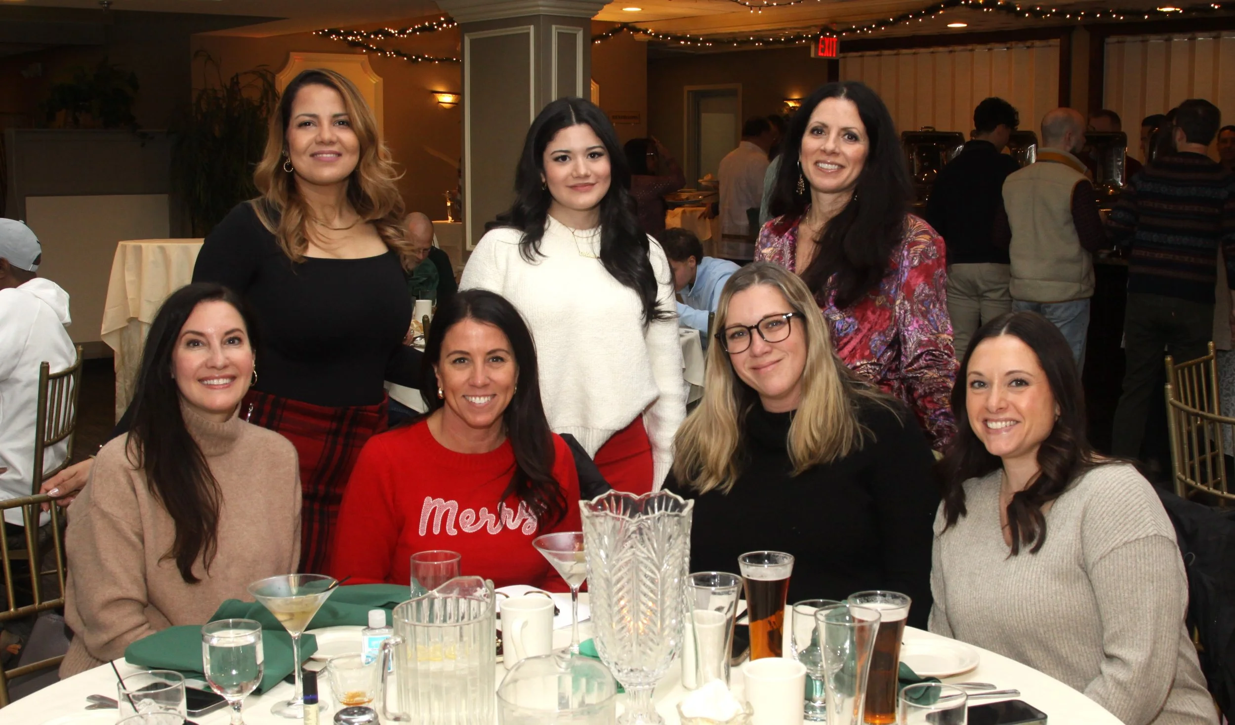 Group of women celebrating holiday dinner at a decorated restaurant, smiling and sitting around a table with drinks and festive decorations.