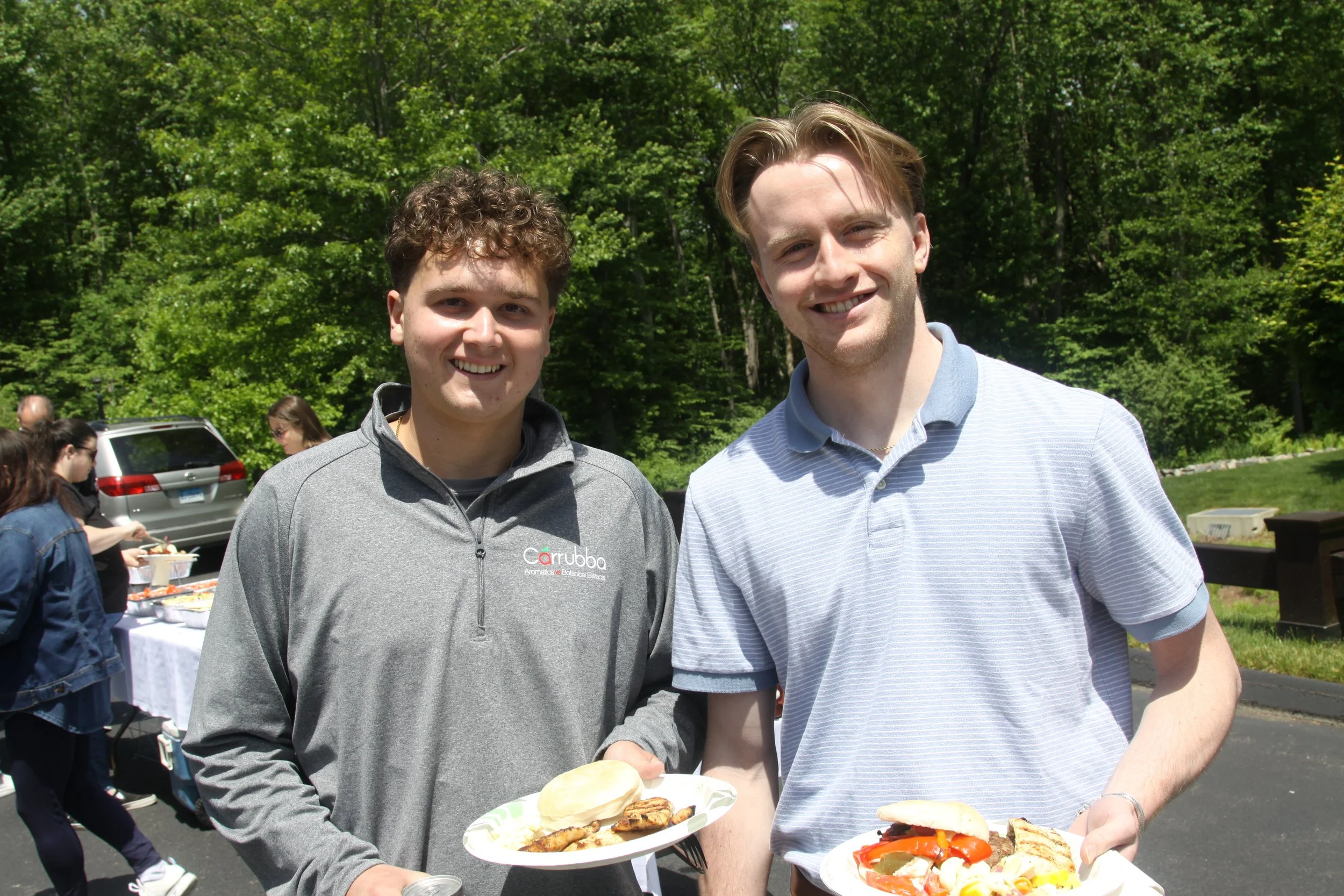 Two young men standing outdoors at a gathering, smiling and holding plates of food including grilled chicken, vegetables, and bread, with green trees and other people in the background.