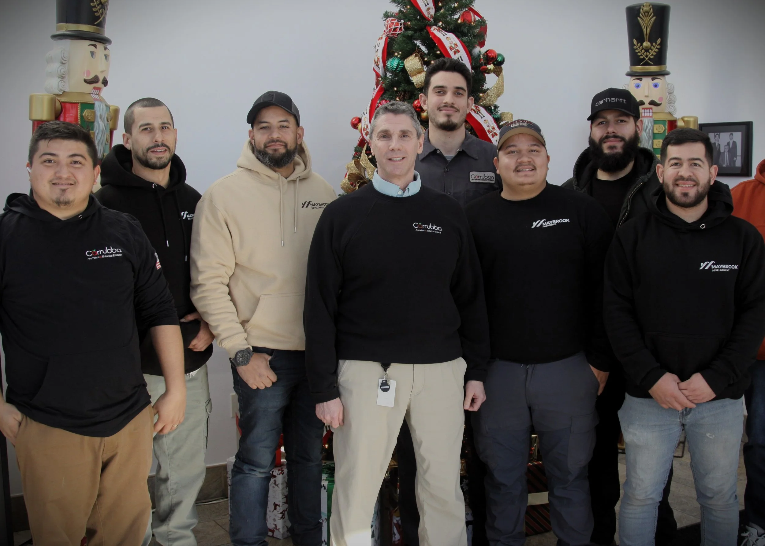 Group of eight men standing in front of a decorated Christmas tree, smiling at the camera. Some are wearing black hoodies with logos, and one man is dressed in a light tan hoodie. The background includes assorted Christmas decorations and tall nutcra