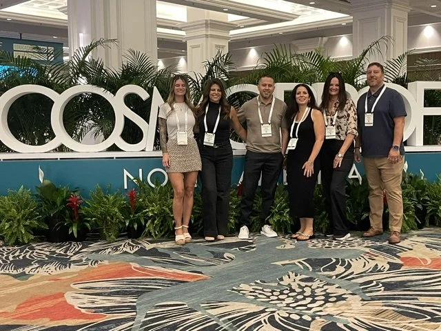 Group of six people standing together indoors at a conference or event, with a large sign reading 'COSM' behind them and decorative plants. They are smiling and wearing name tags.