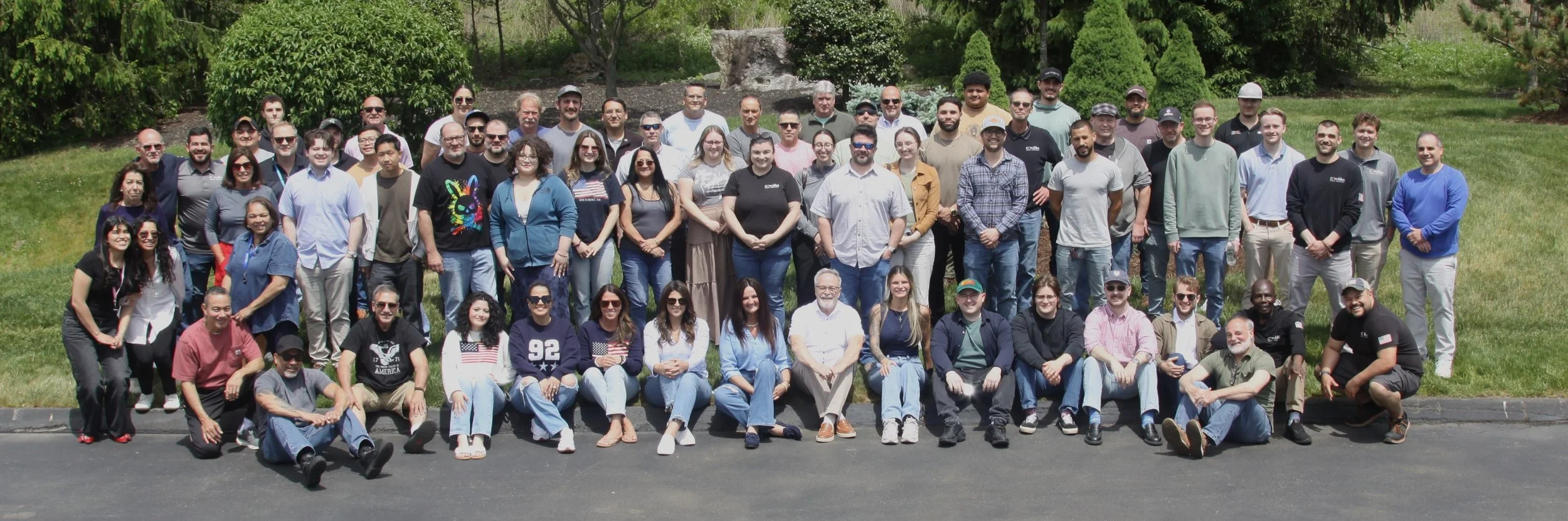 Group photo of numerous people standing and sitting outdoors on a grassy area with trees in the background.