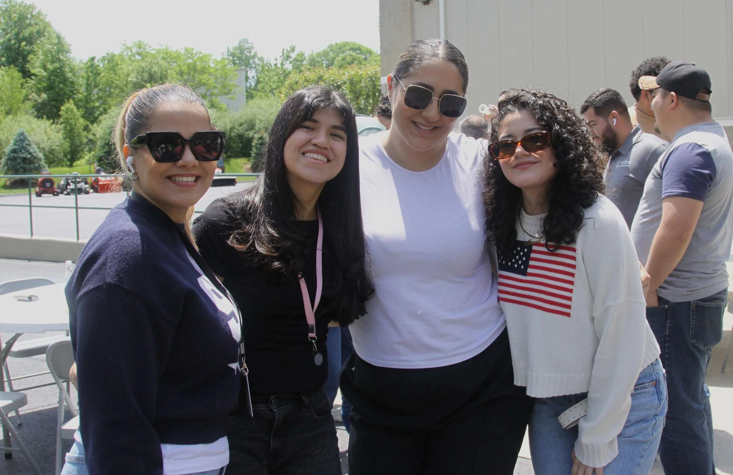 Group of four women smiling at an outdoor event, wearing sunglasses, with other people and green trees in the background.