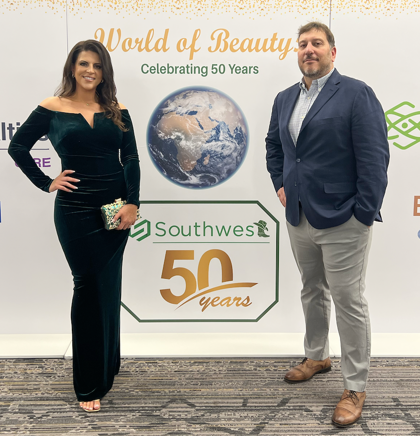 Two people, a woman in a black velvet dress and a man in a navy blazer and light gray pants, standing in front of a backdrop celebrating Southwesto's 50 years. The backdrop features a globe and text that says 'World of Beauty' and 'Celebrating 50 Yea