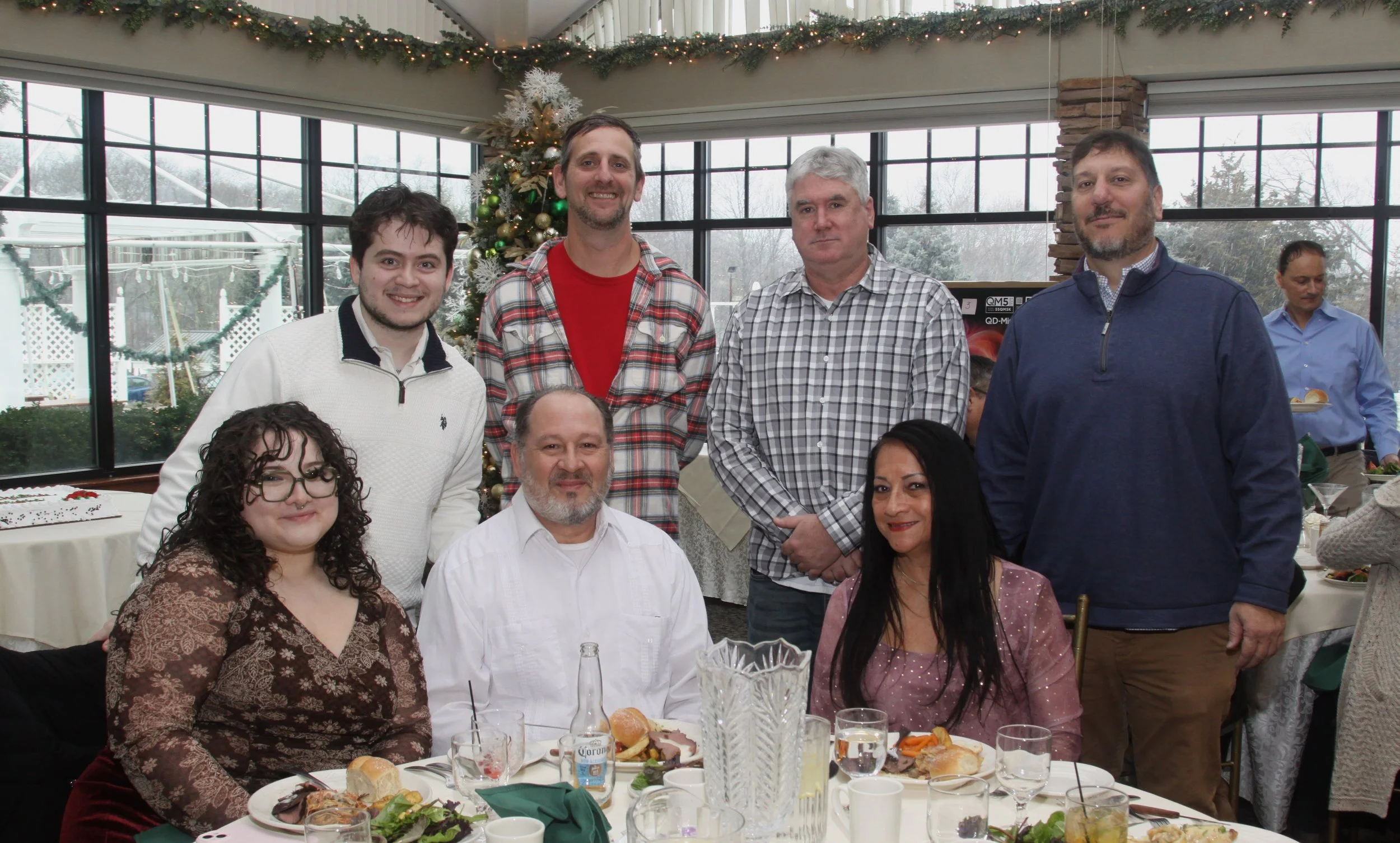 A group of people gathered for a holiday or Christmas celebration indoors, with a decorated Christmas tree in the background, large windows with snowy outdoor scenery, and a table set with food and drinks.