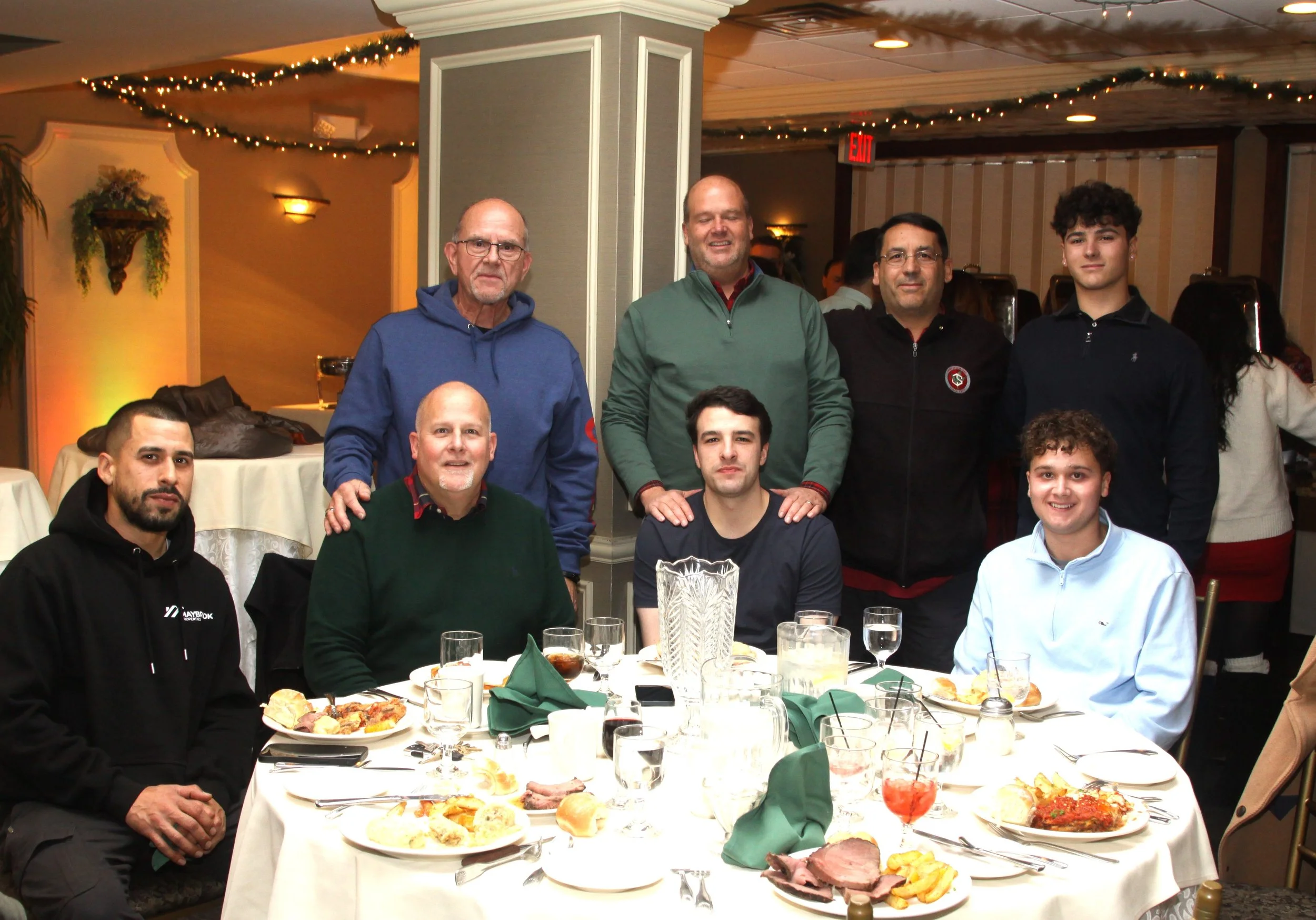 A group of people gathered around a table at a festive dinner event, with food, drinks, and holiday decorations in the background.