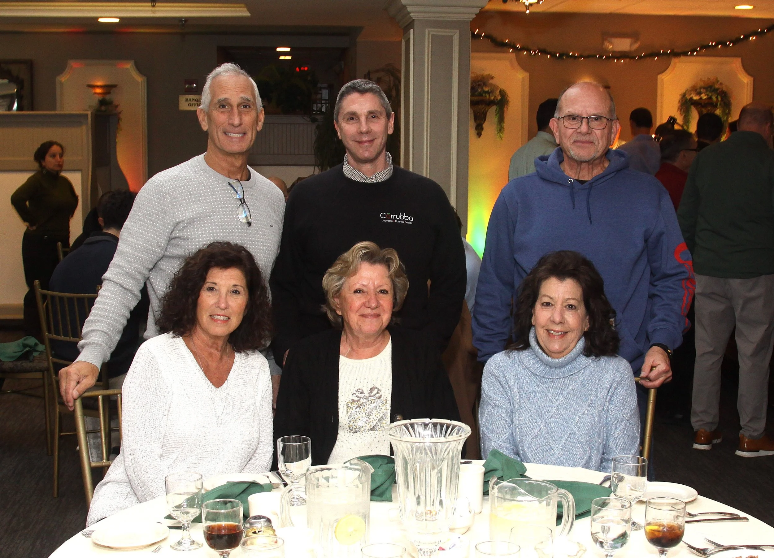 Group of people at a dinner event, sitting and standing around a table with glasses and dishes, in a decorated banquet hall.