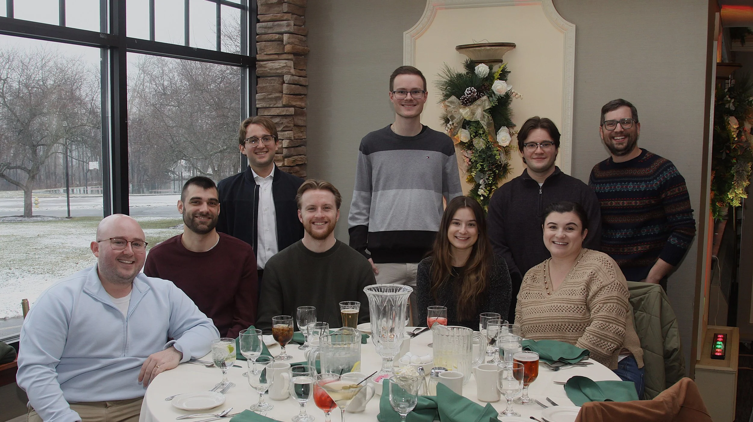 Group of nine people gathered around a dining table with plates, glasses, and drinks, indoors near large windows with a winter landscape outside. A decorative Christmas wreath hangs on the wall behind them.