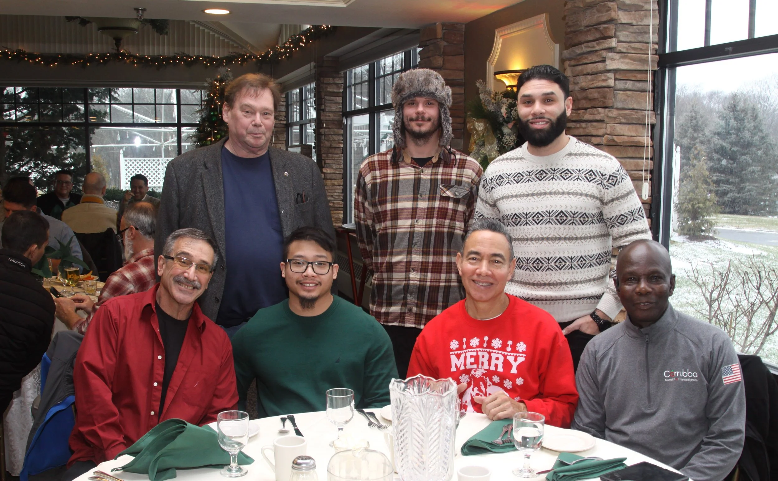 Group of men gathered for a festive holiday meal indoors, with holiday decorations and large windows in the background.