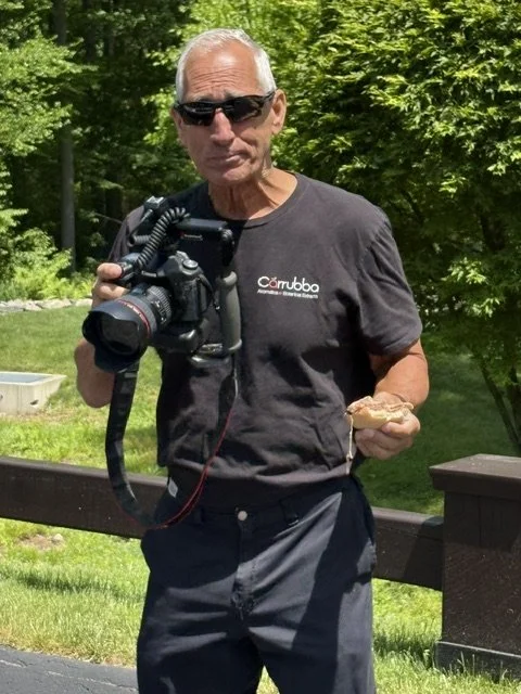 Older man with sunglasses holding a professional camera in one hand and a sandwich in the other, standing outdoors in a park with green trees and grass.