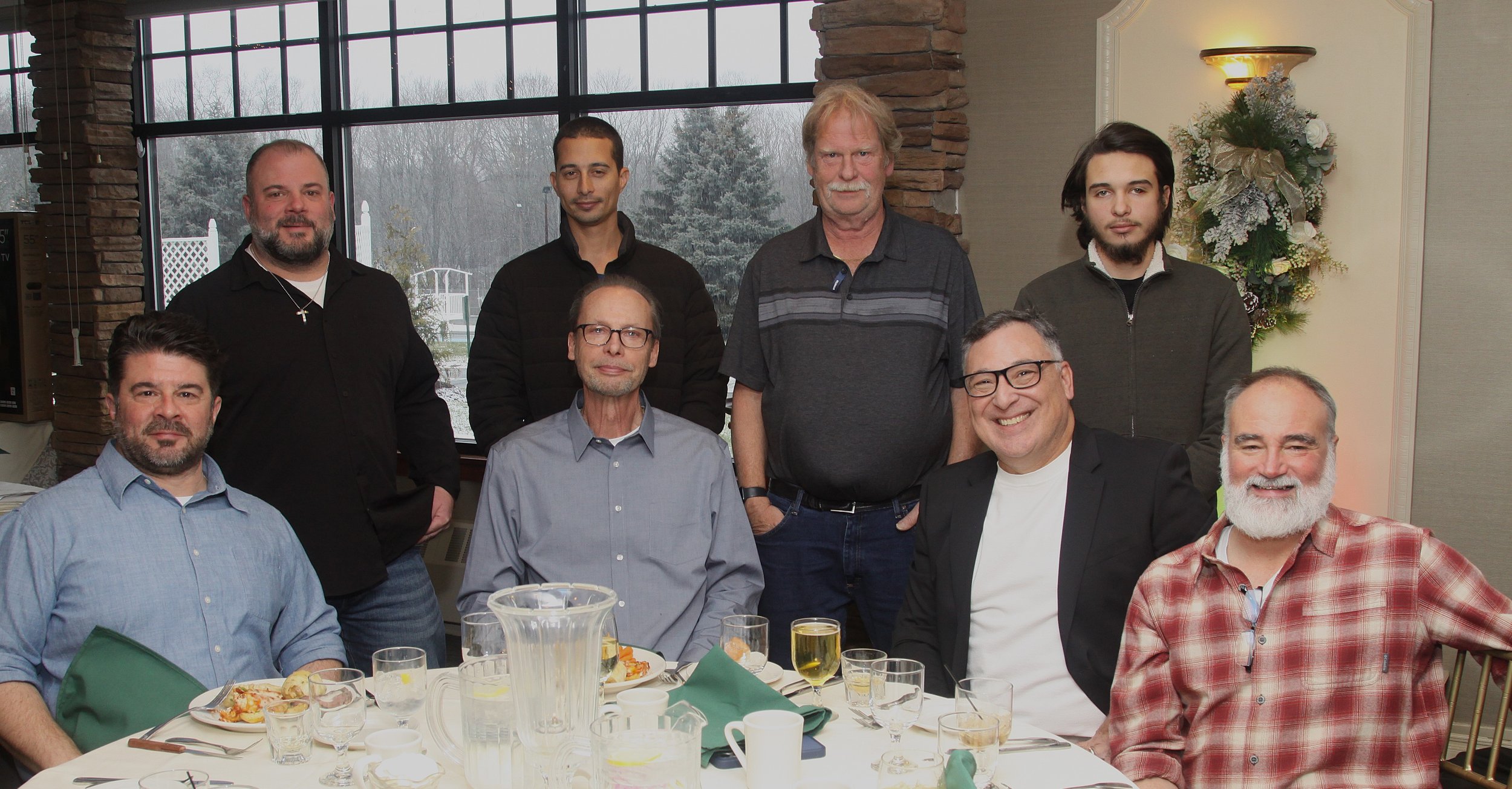 A group of ten men gathered in a restaurant or banquet room, with large windows showing a snowy outdoor scene. They are sitting and standing around a table set with plates of food, drinks, and tableware, smiling and posing for the photo.