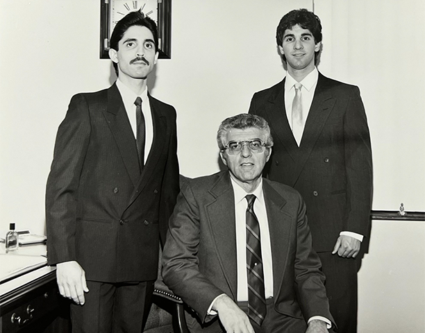 Three men in formal suits and ties posing for a black-and-white photograph indoors, with one seated in the center and two standing behind him, against a plain wall with a clock.