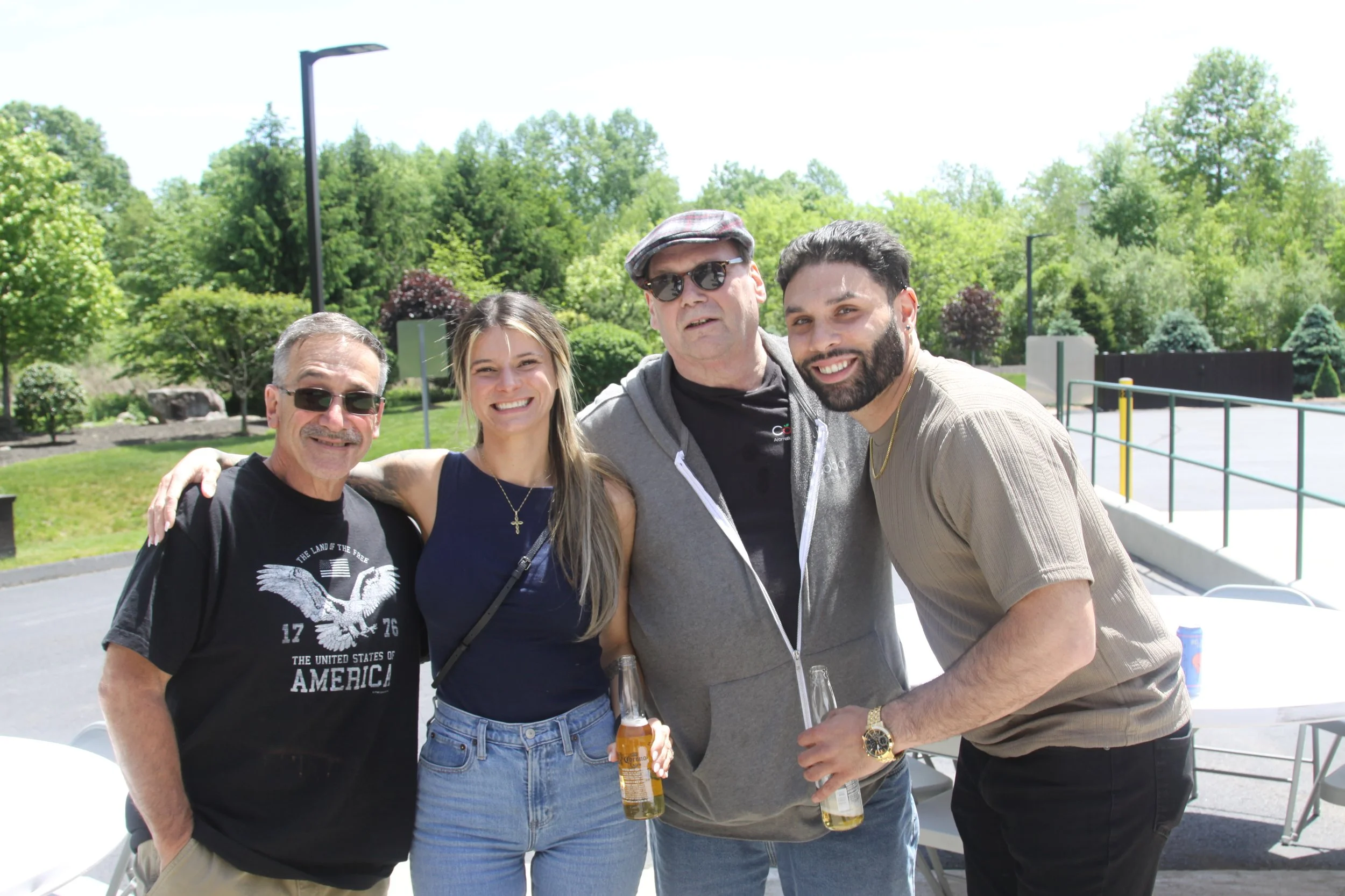 Four people standing outdoors smiling and posing for a photo, with green trees and a blue sky in the background.
