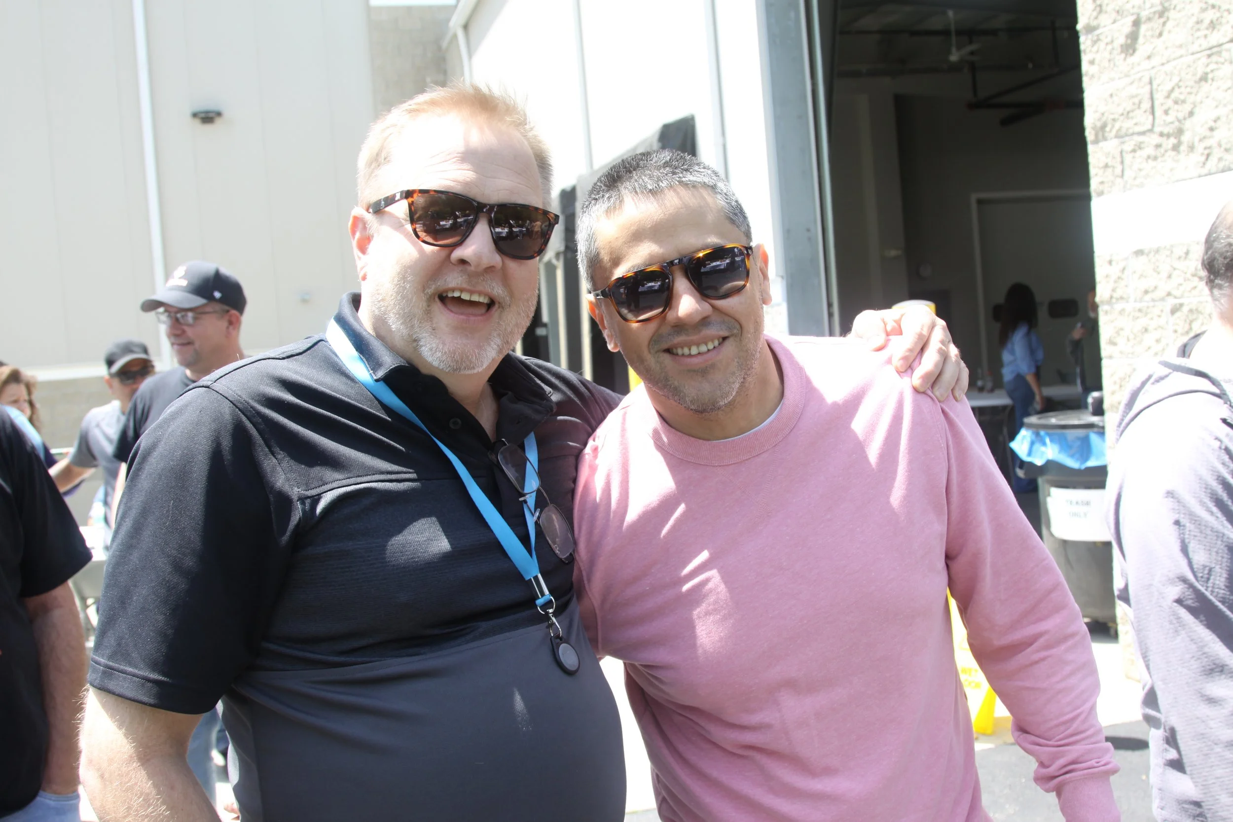 Two men smiling and wearing sunglasses at an outdoor event, with other people in the background.