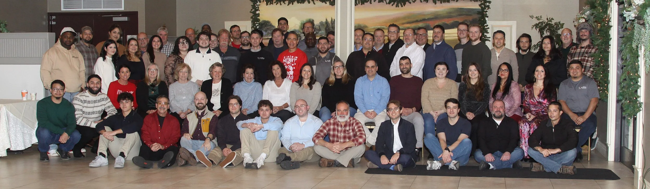 A large group of people gathered indoors for a group photo, with some people sitting on the floor and others standing behind them, in a decorated room with a landscape painting and greenery in the background.