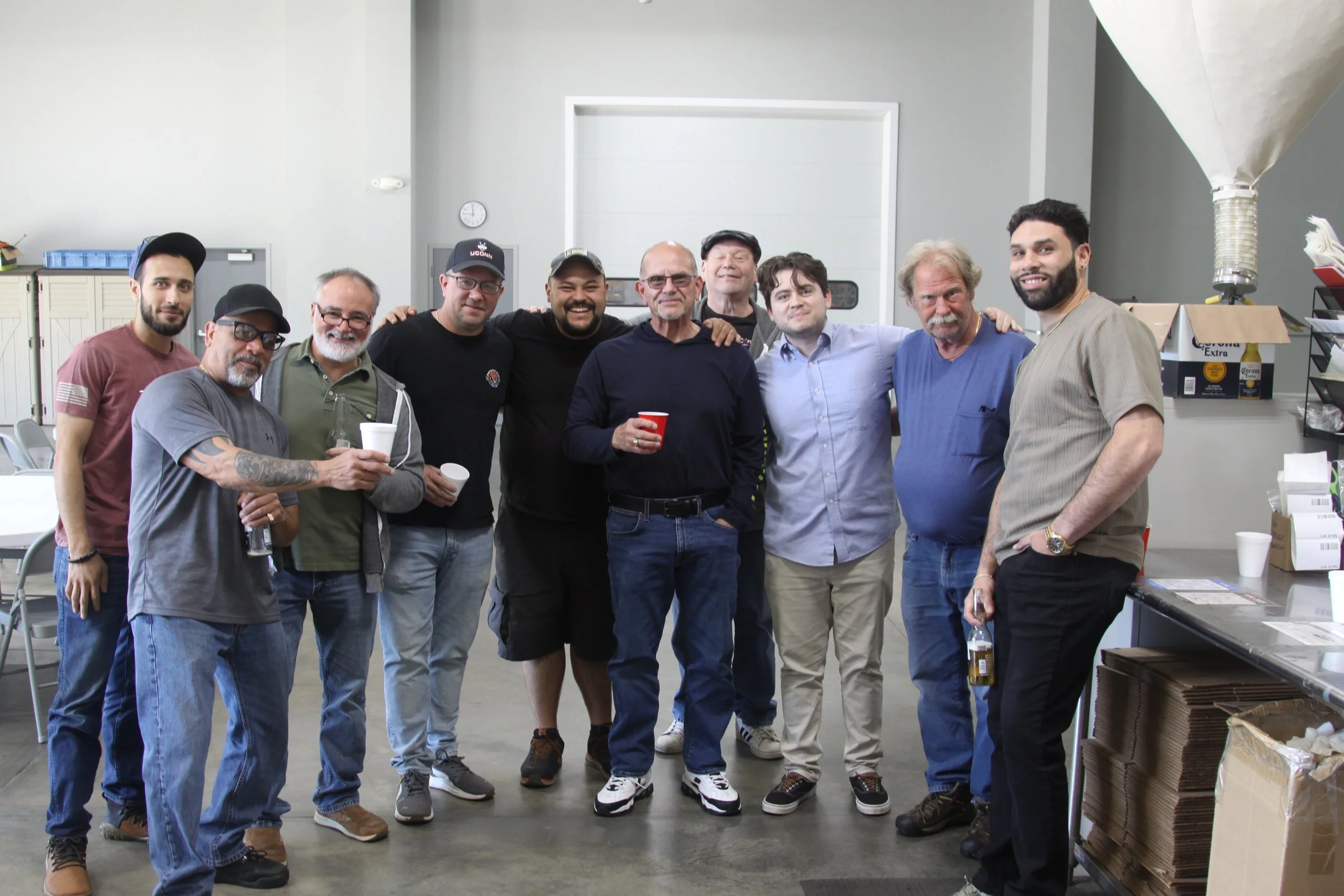 A group of eleven men standing closely together in an indoor space, smiling at the camera. They are dressed casually, with some holding drinks. The background shows industrial storage shelves, boxes, and equipment.