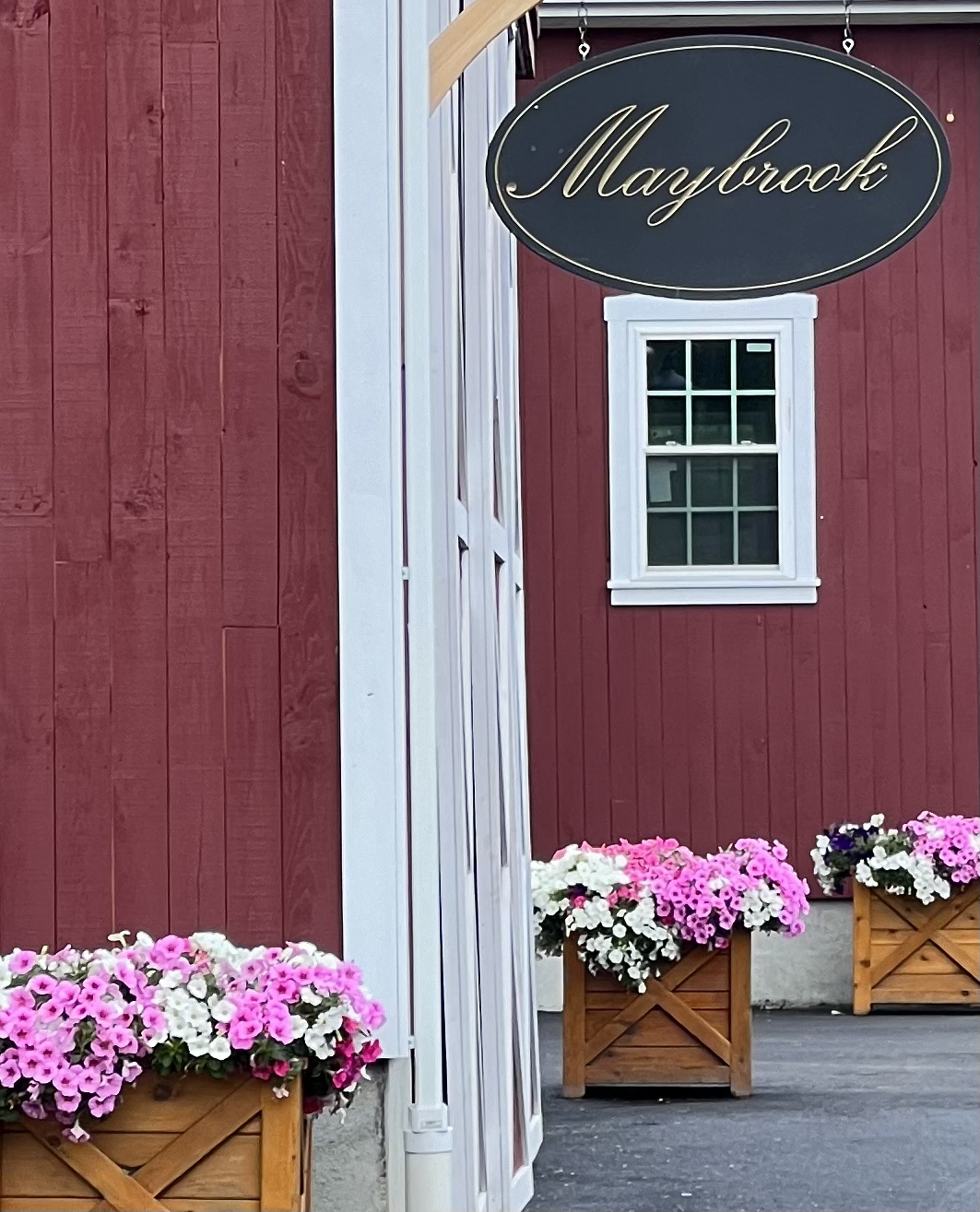 Red wooden building with a white-framed window, hanging oval sign with the word 'Maybrock' in cursive, and pink and white flowers in wooden planters outside.