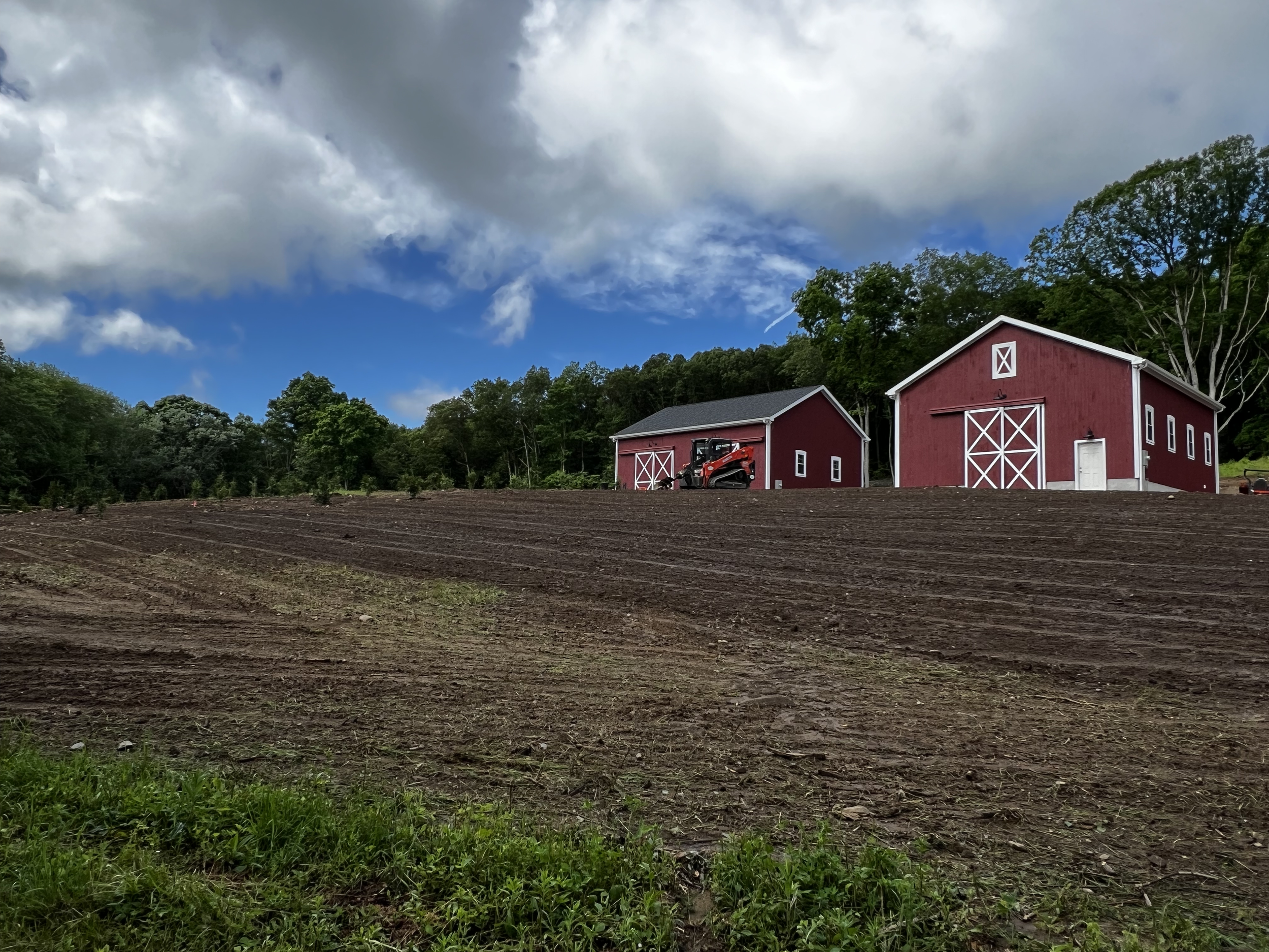 A rural landscape with two red barns, a patch of green grass, tilled soil, a partly cloudy sky, and surrounding green trees.