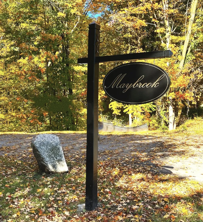 A black signpost with an oval sign reading 'Mayblook' hangs from a horizontal arm in an outdoor setting during fall, with colorful autumn leaves and trees in the background.
