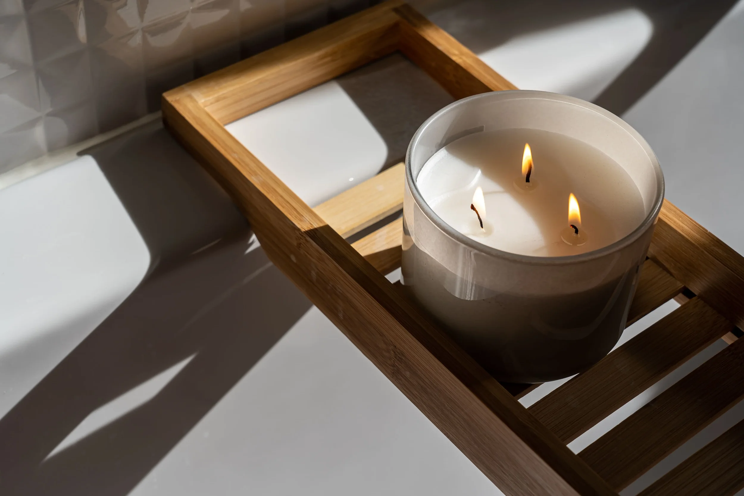 A lit white candle inside a glass container on a wooden tray, casting shadows on a tiled wall.