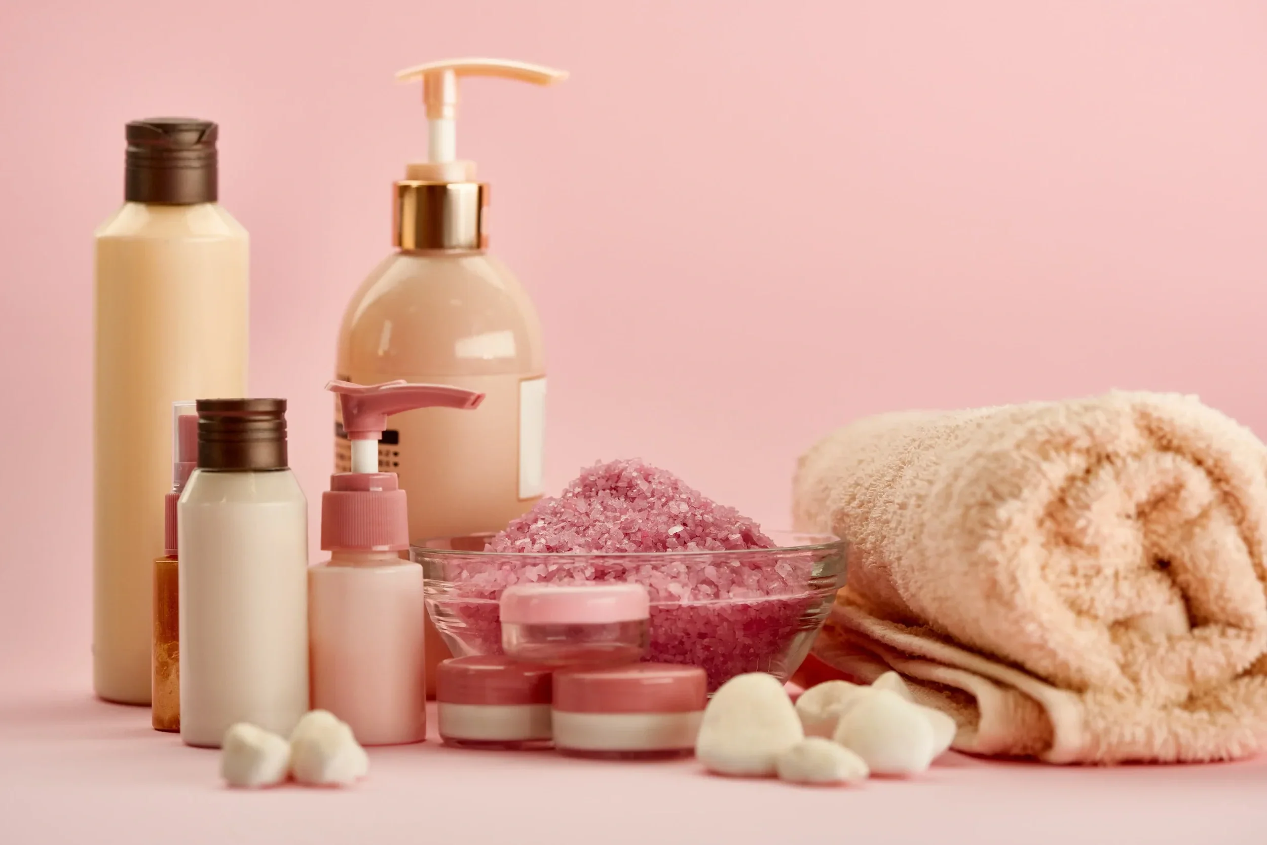 Various skincare bottles and jars, pink bath salts in a glass bowl, a rolled towel, and white bath bombs against a pink background.